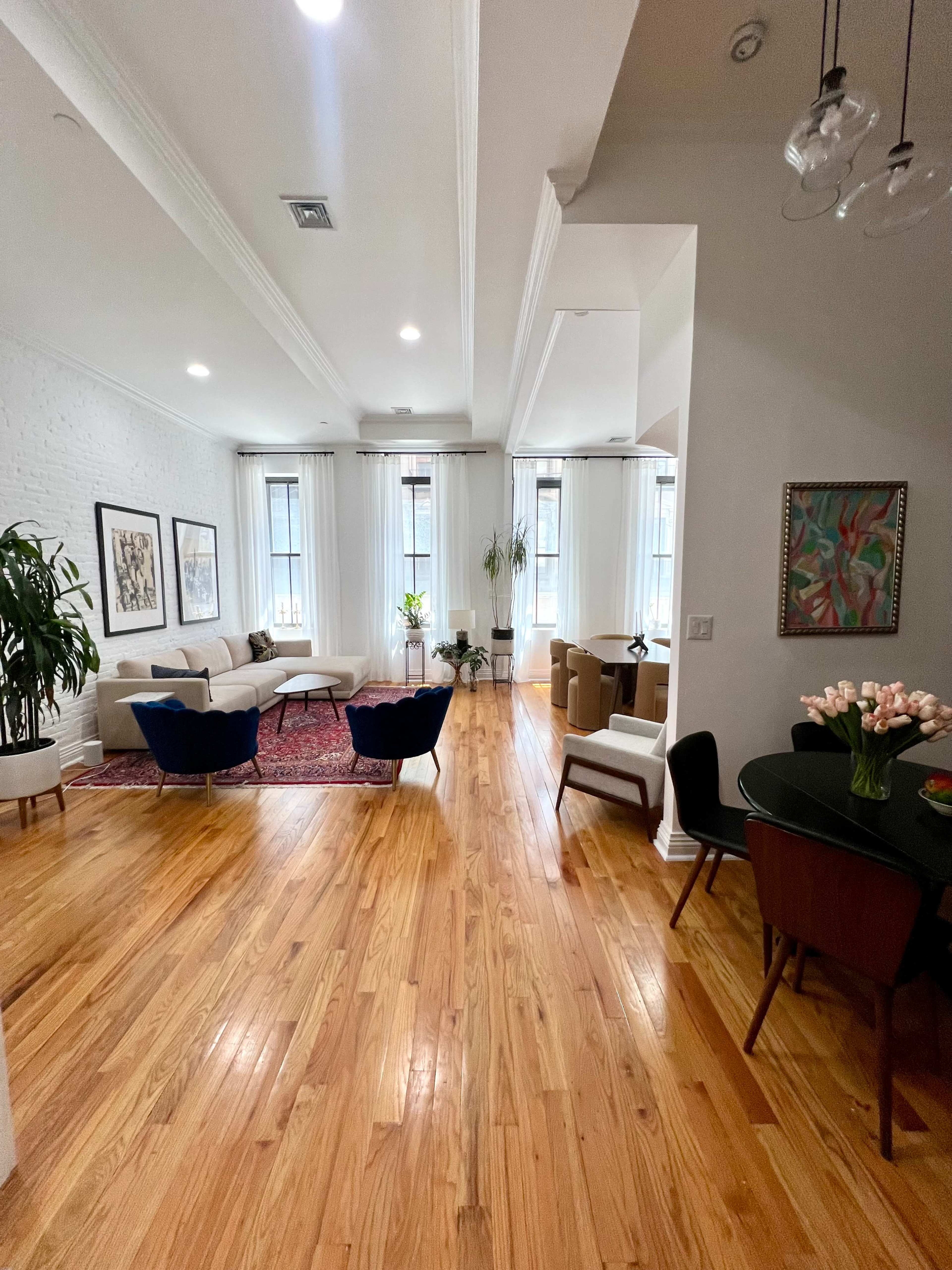 A spacious living area features hardwood floors, large windows with sheer curtains, and a variety of seating arrangements including a dining table, sofas, and armchairs.