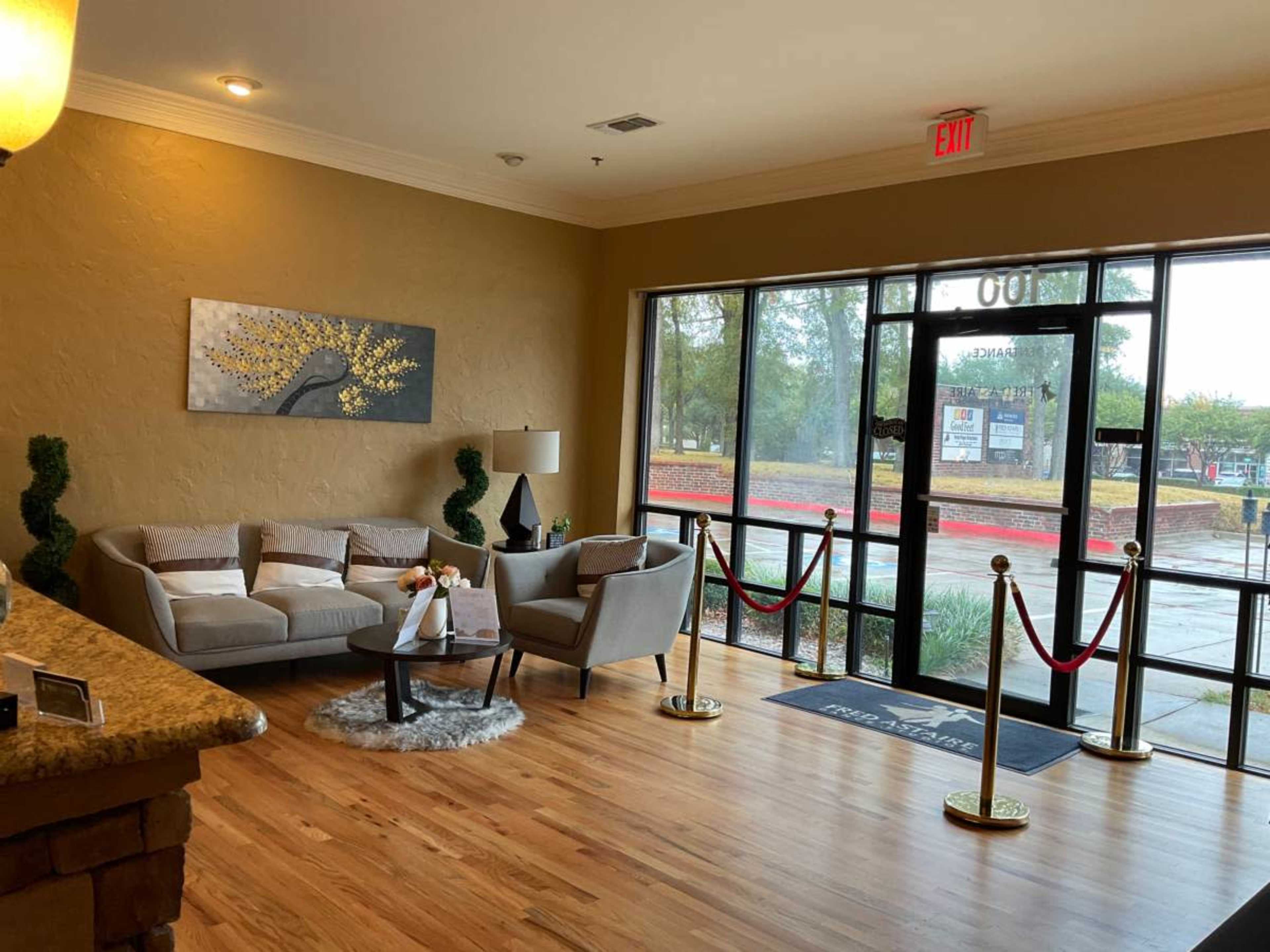 The image shows a reception area with a light-colored wall, a comfortable seating arrangement, and large windows providing a view of the outside.