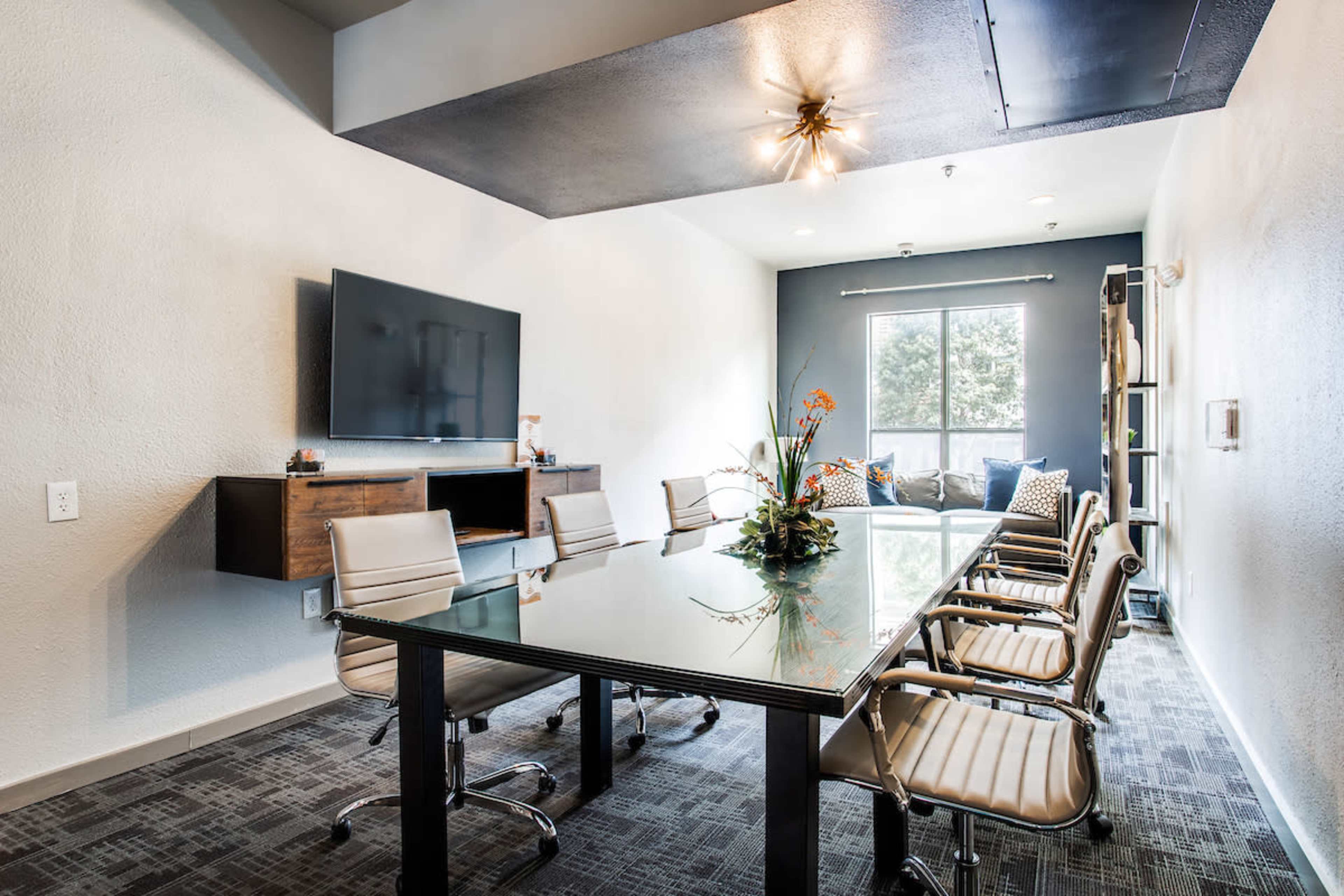 A modern conference room features a glass table surrounded by eight chrome chairs, with a large television mounted on the wall and a seating area in the background.