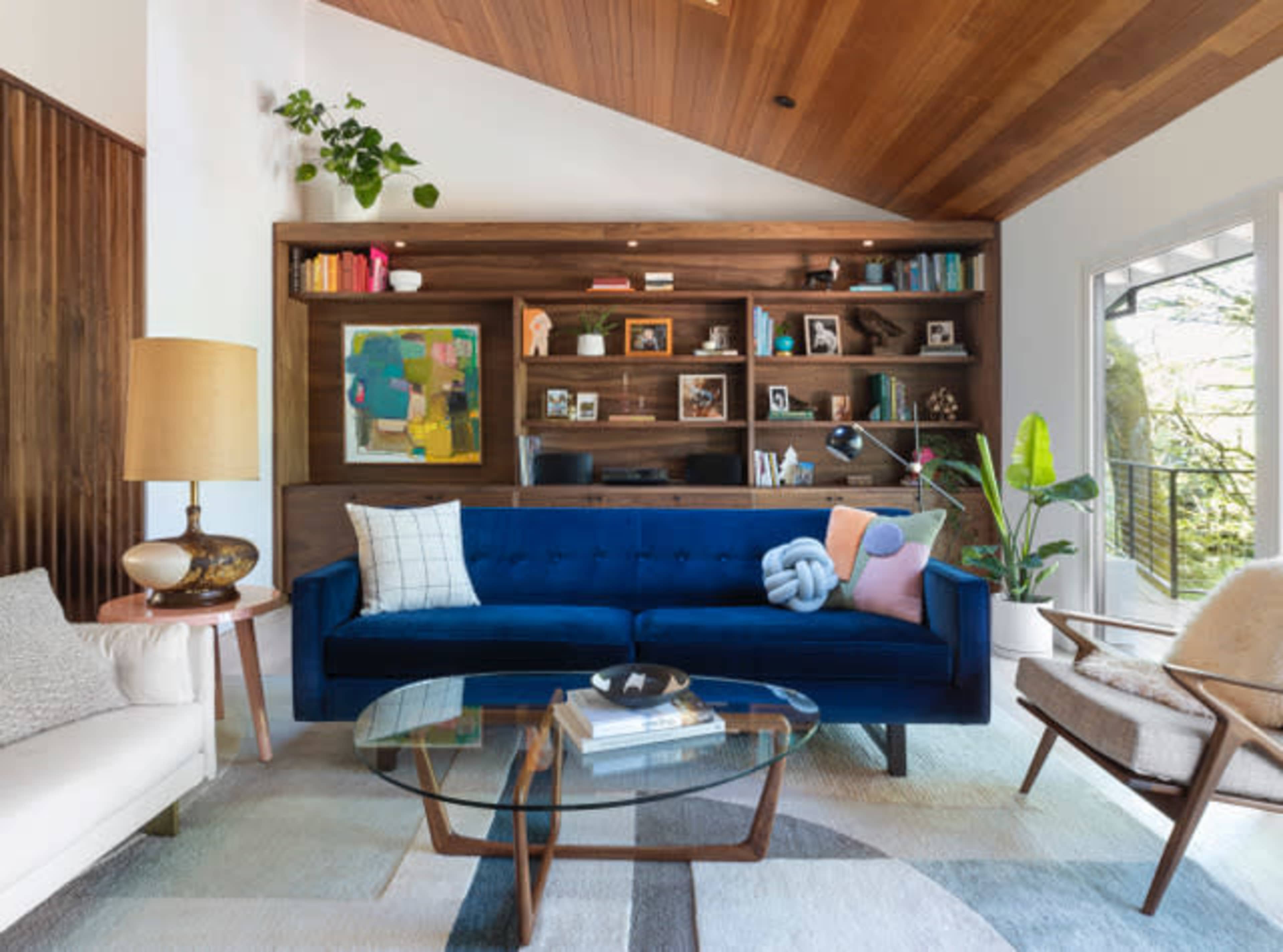 A modern living room featuring a blue velvet sofa, a glass coffee table, and a wooden bookshelf filled with books and decorative items.
