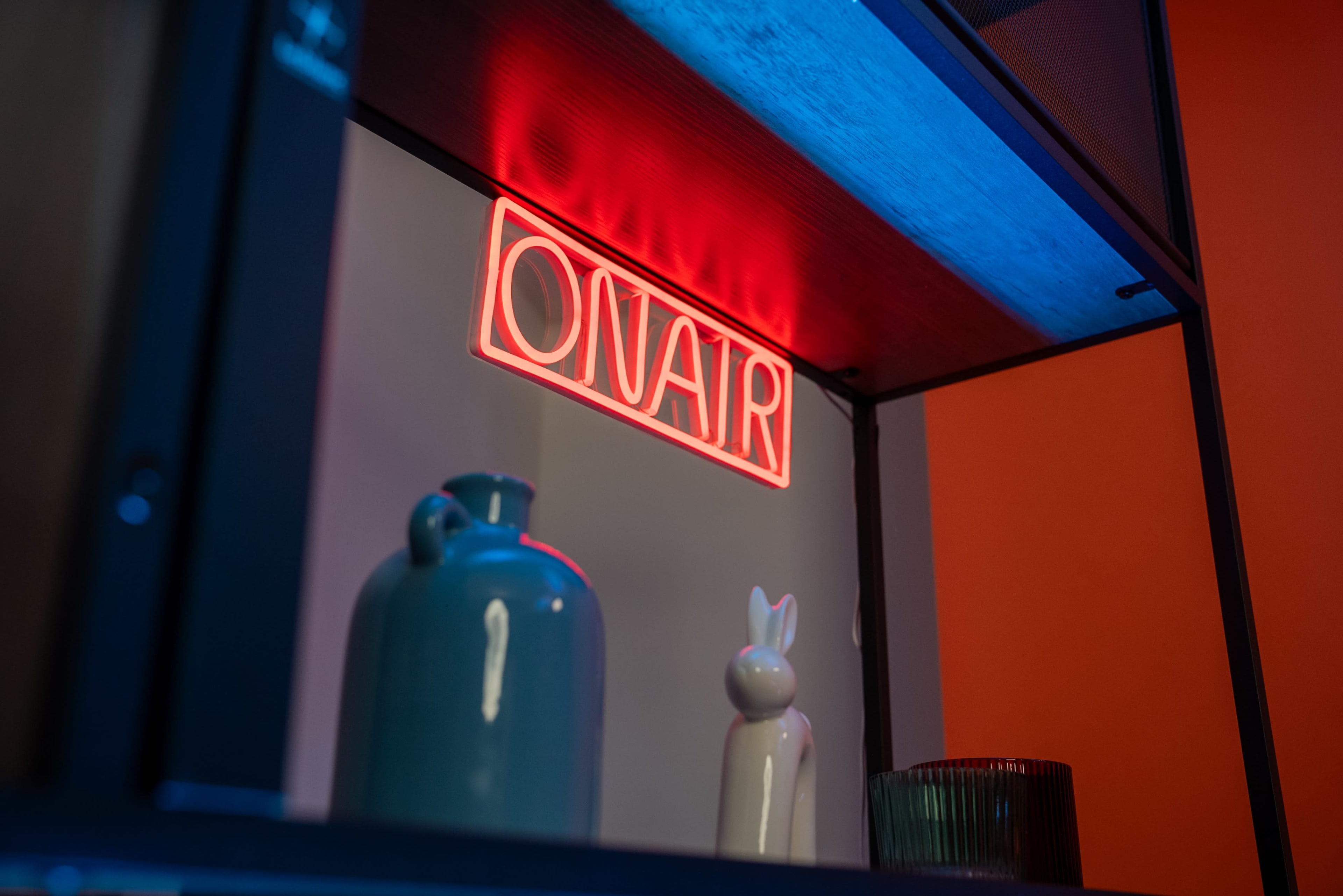A brightly lit "ON AIR" sign is displayed on a wall shelf alongside a blue vase, a white rabbit figurine, and a glass container.