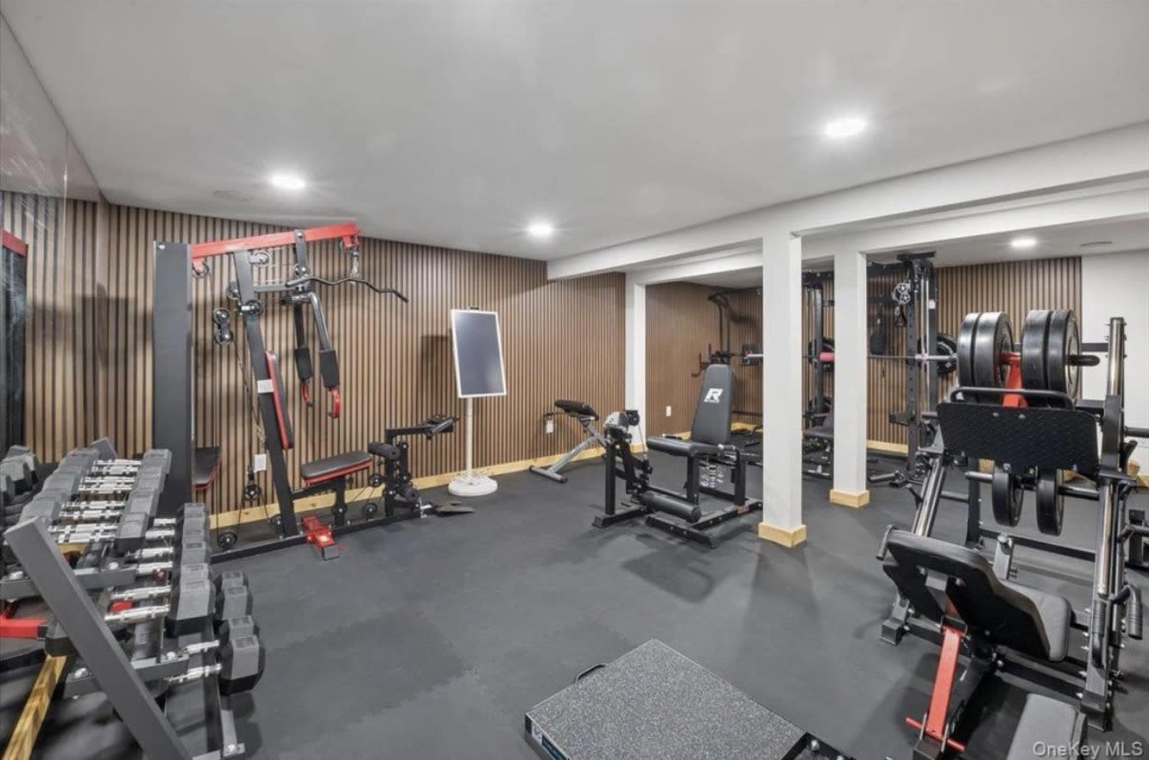 The image shows a home gym equipped with various weightlifting and exercise machines, along with free weights and workout benches.