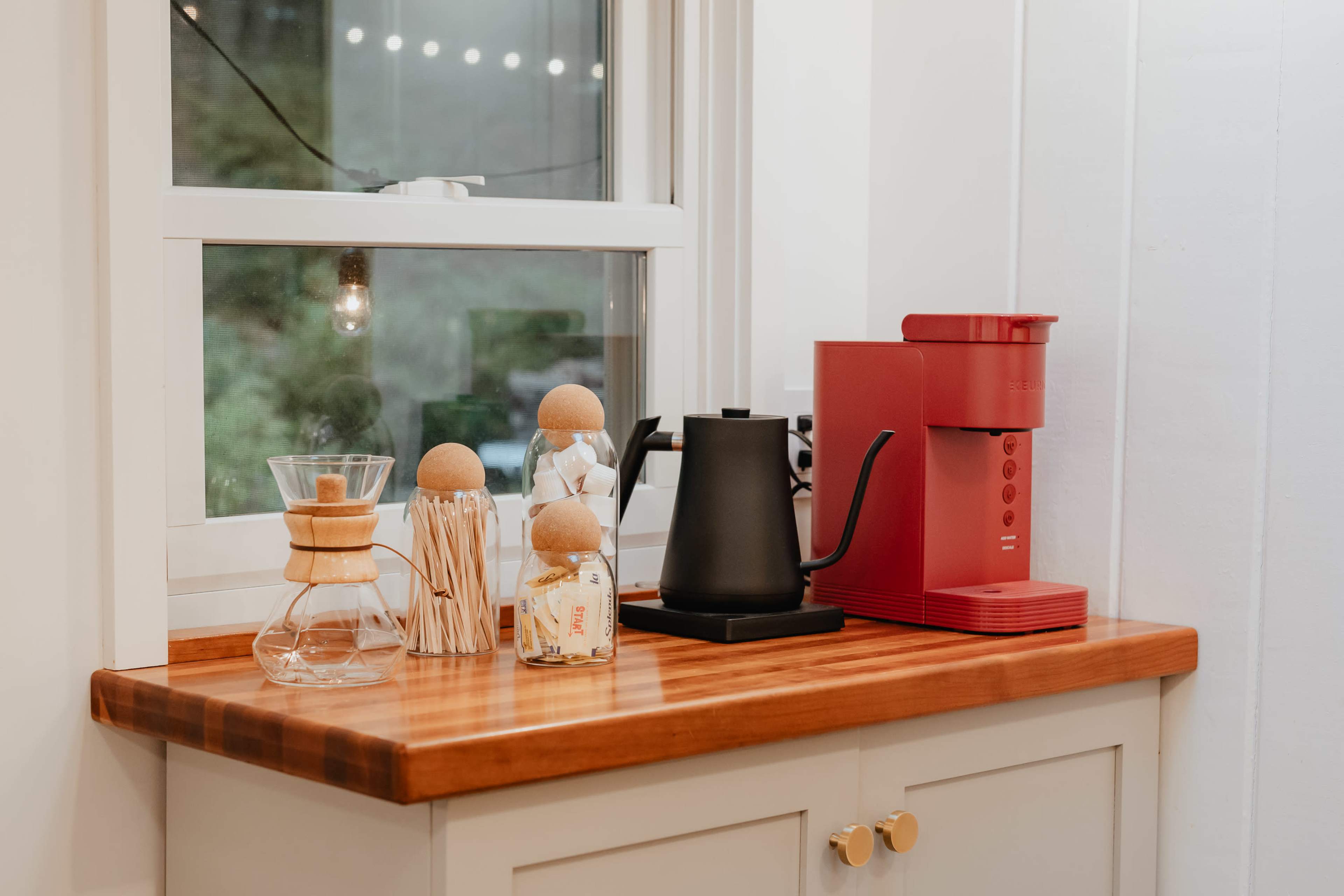 A wooden countertop holds a red coffee machine, a black kettle, a glass coffee maker, and decorative items near a window.