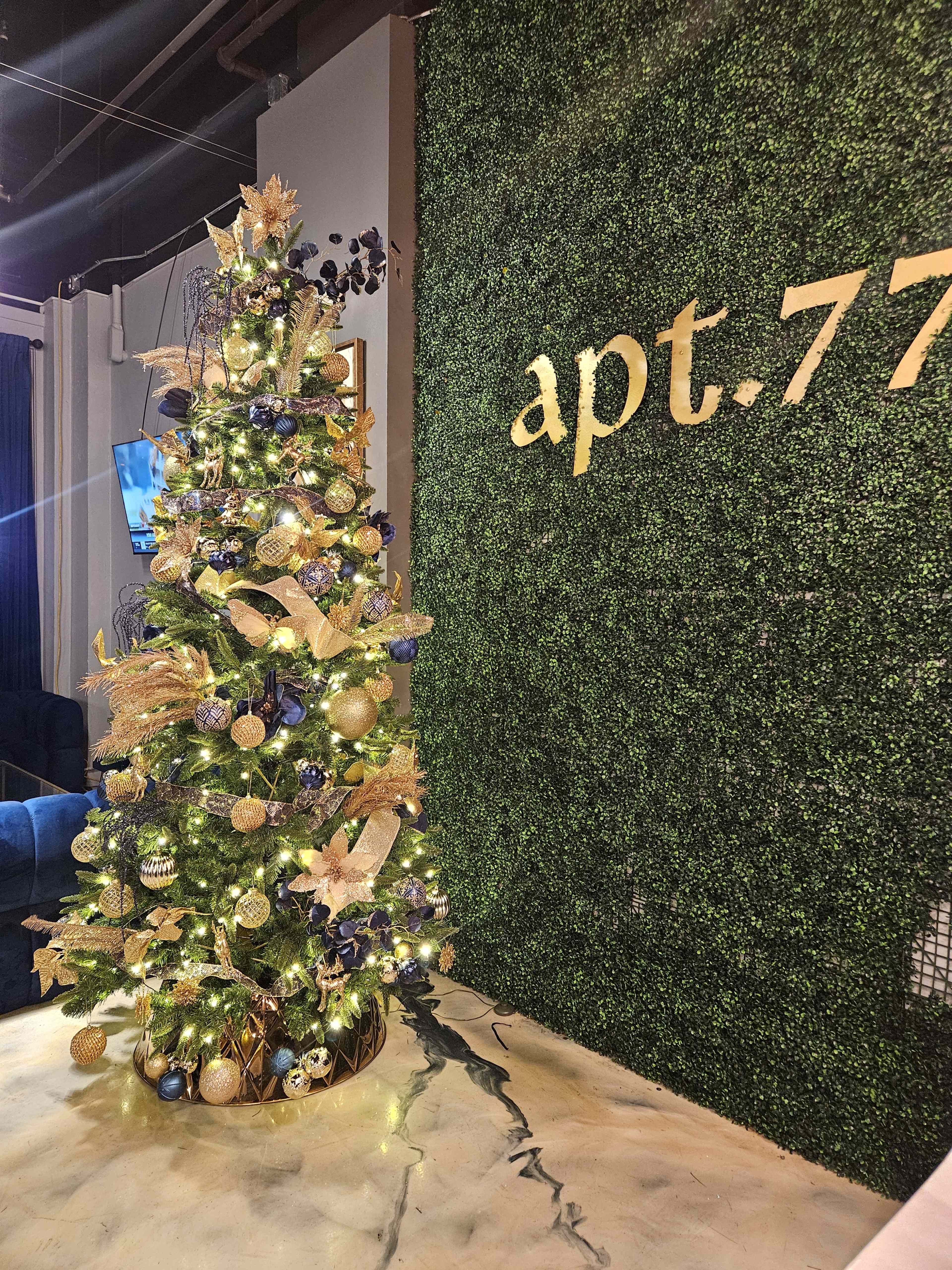 A decorated Christmas tree with gold and blue ornaments is positioned next to a green wall featuring the text "apt.77."