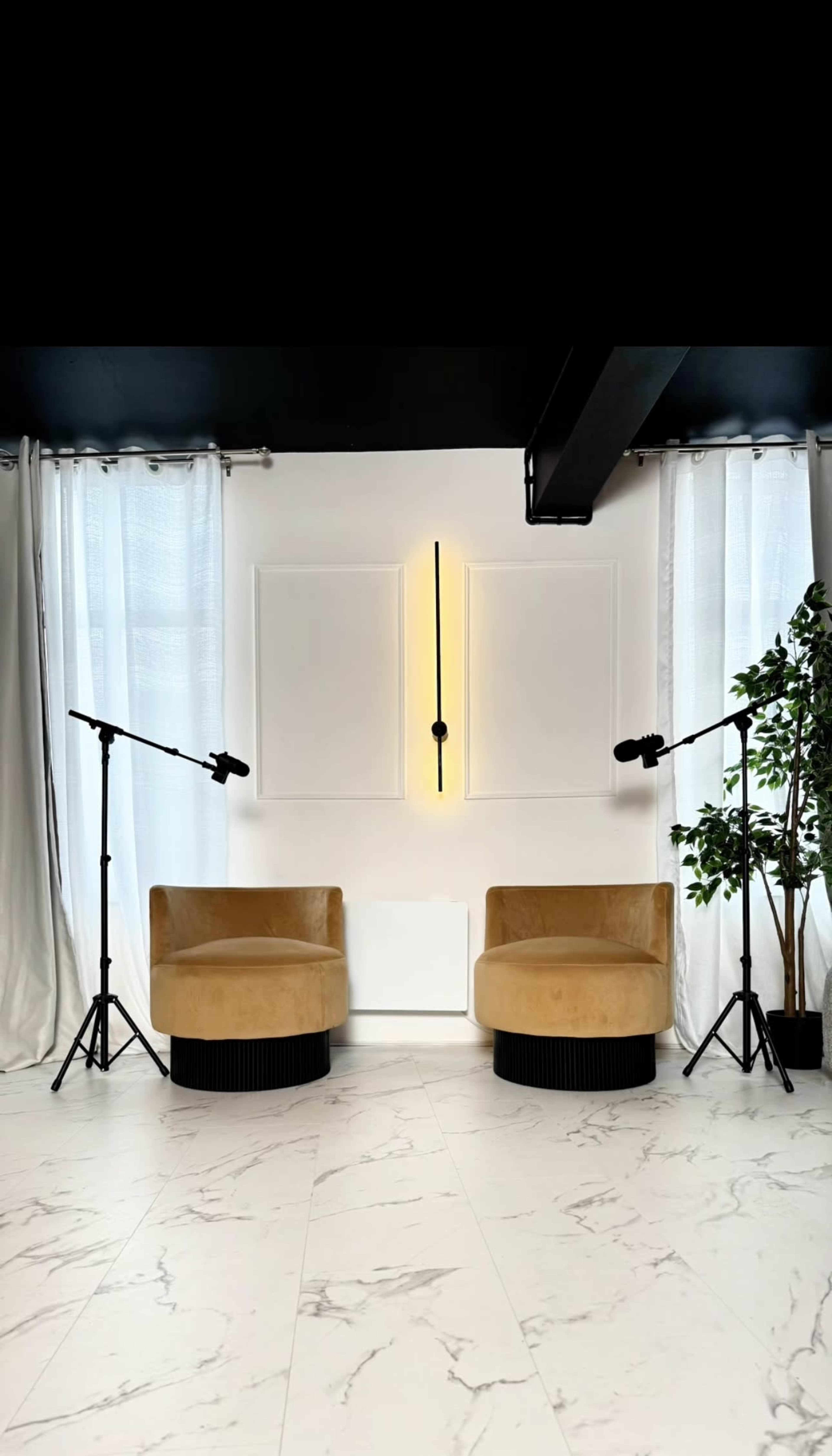 The image shows a modern interior space featuring two yellow chairs positioned across from a white wall with a vertical light fixture, flanked by two standing microphones and a potted plant.