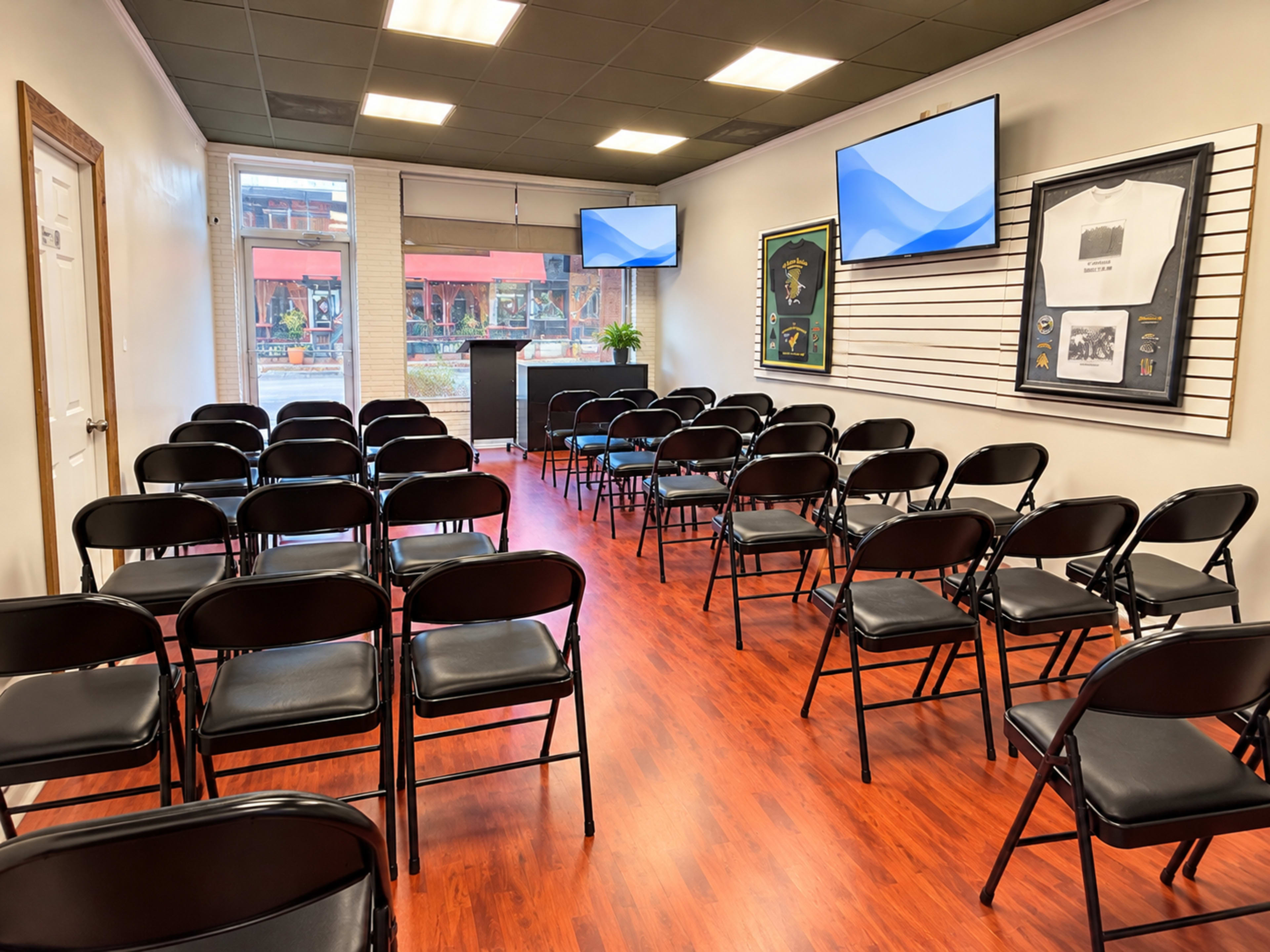 The image shows a neatly arranged room with rows of black folding chairs facing two mounted screens and a podium, with natural light visible through a window.