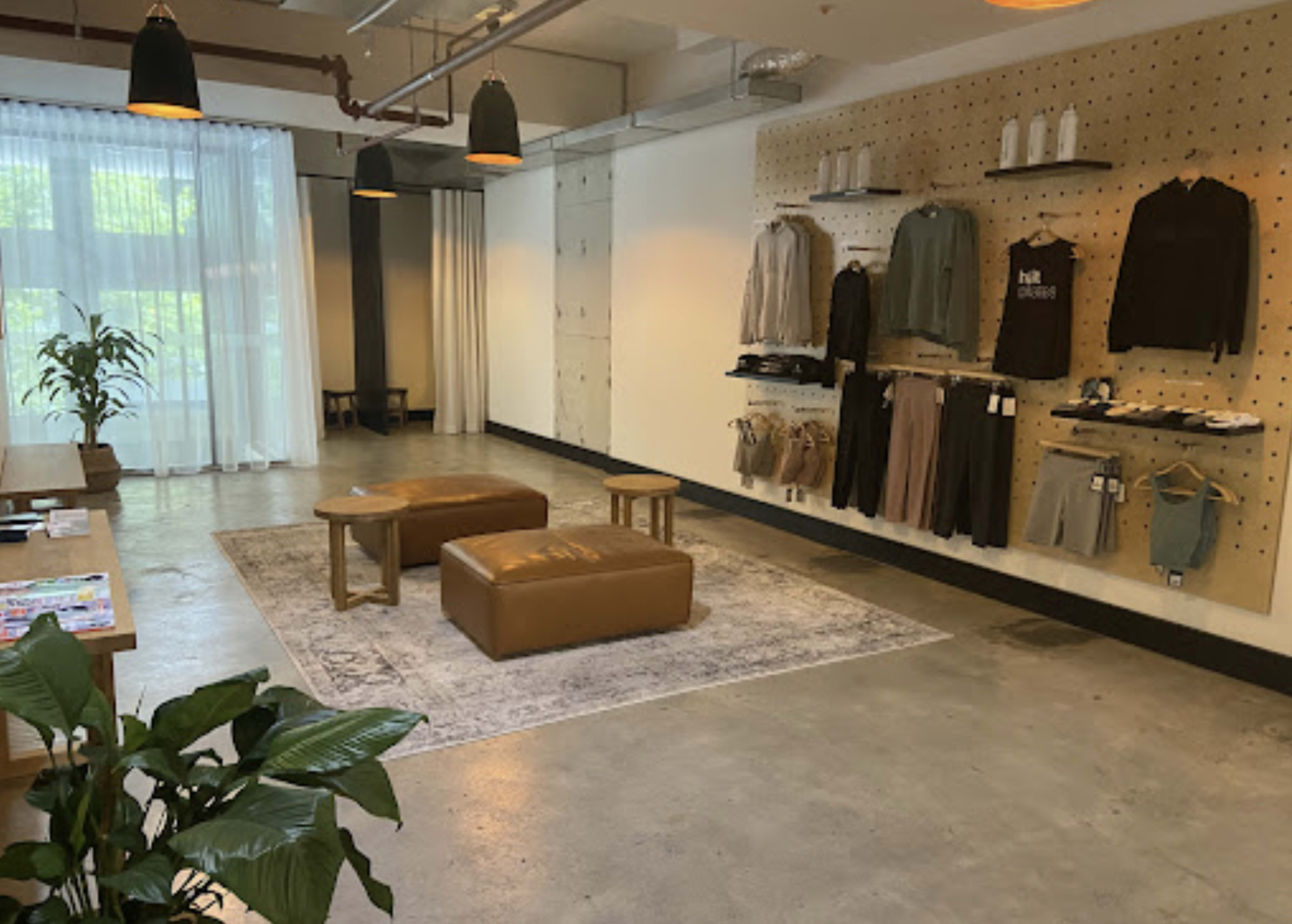 A minimalist retail space featuring a wall of clothing hung on pegs, two leather stools on a rug, and a plant in the foreground.