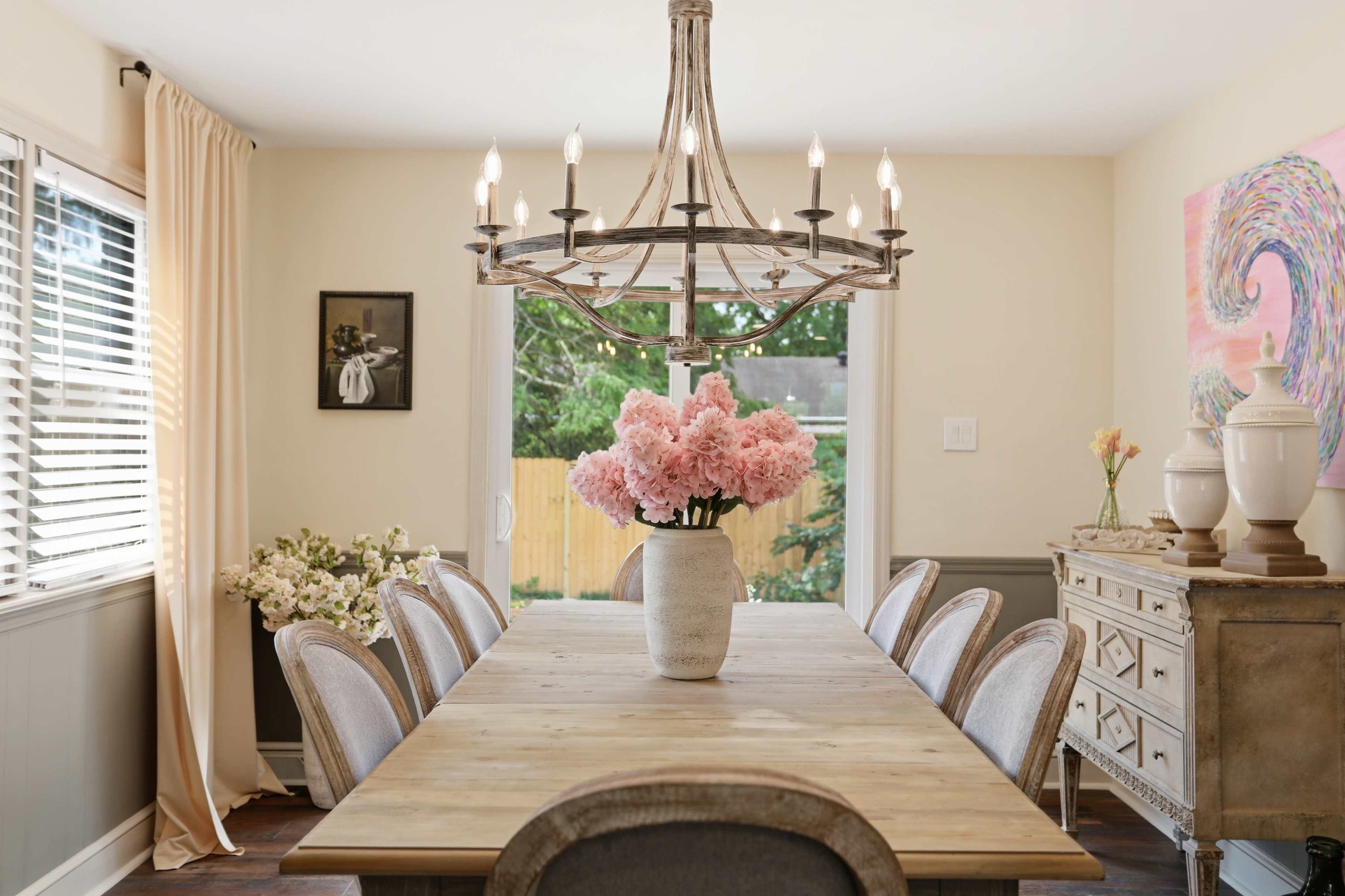 A spacious dining room features a large wooden table surrounded by upholstered chairs, with a chandelier overhead and a vase of pink flowers as a centerpiece.