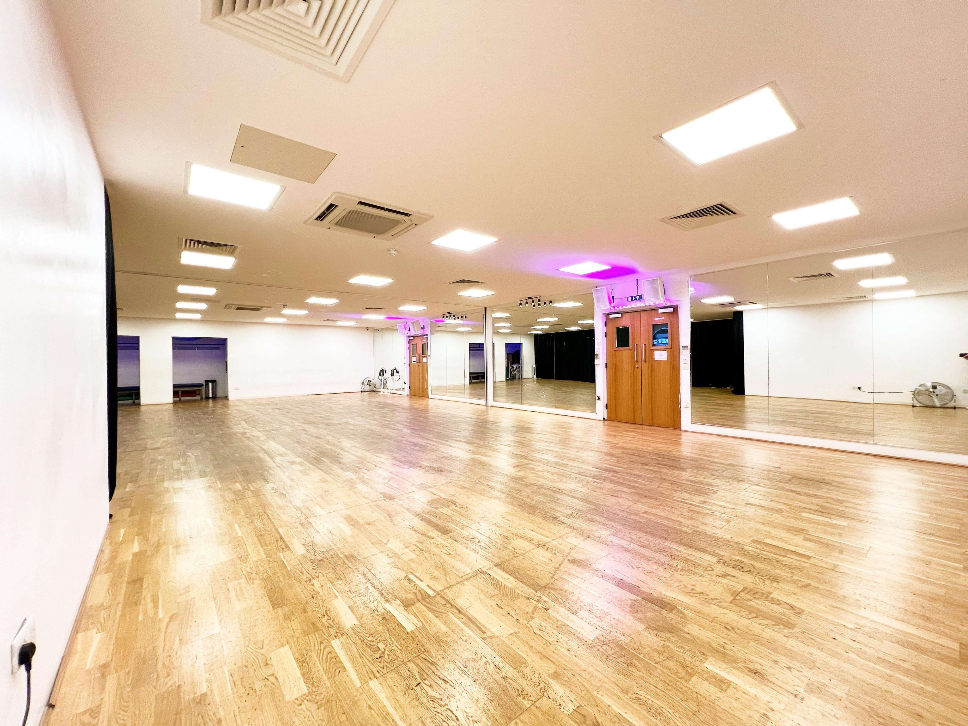 The image shows a spacious dance studio with wooden flooring, bright overhead lights, and large mirrors on one wall.
