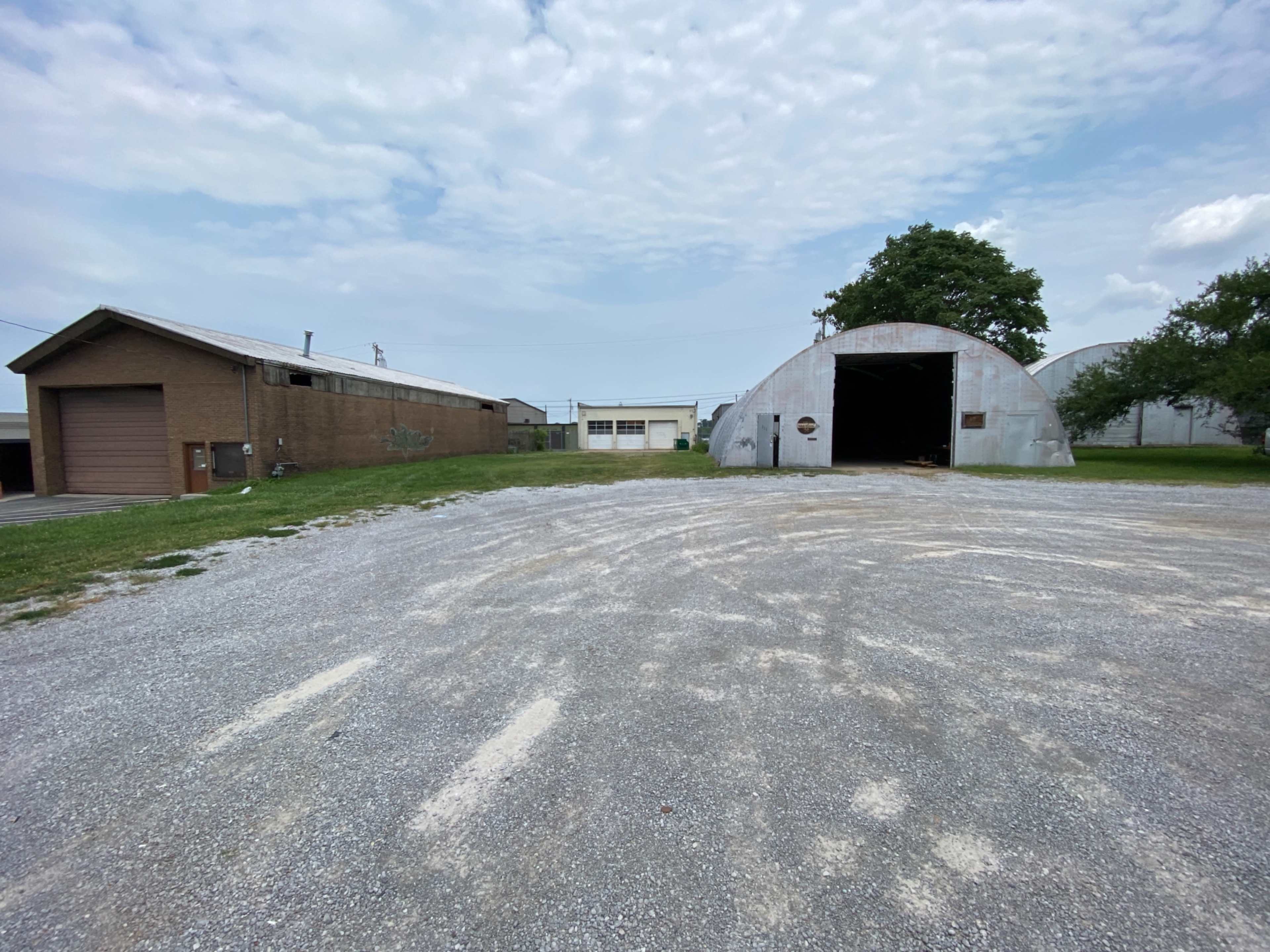 A gravel area with a large, curved metal building and a single-story brick structure on the left.