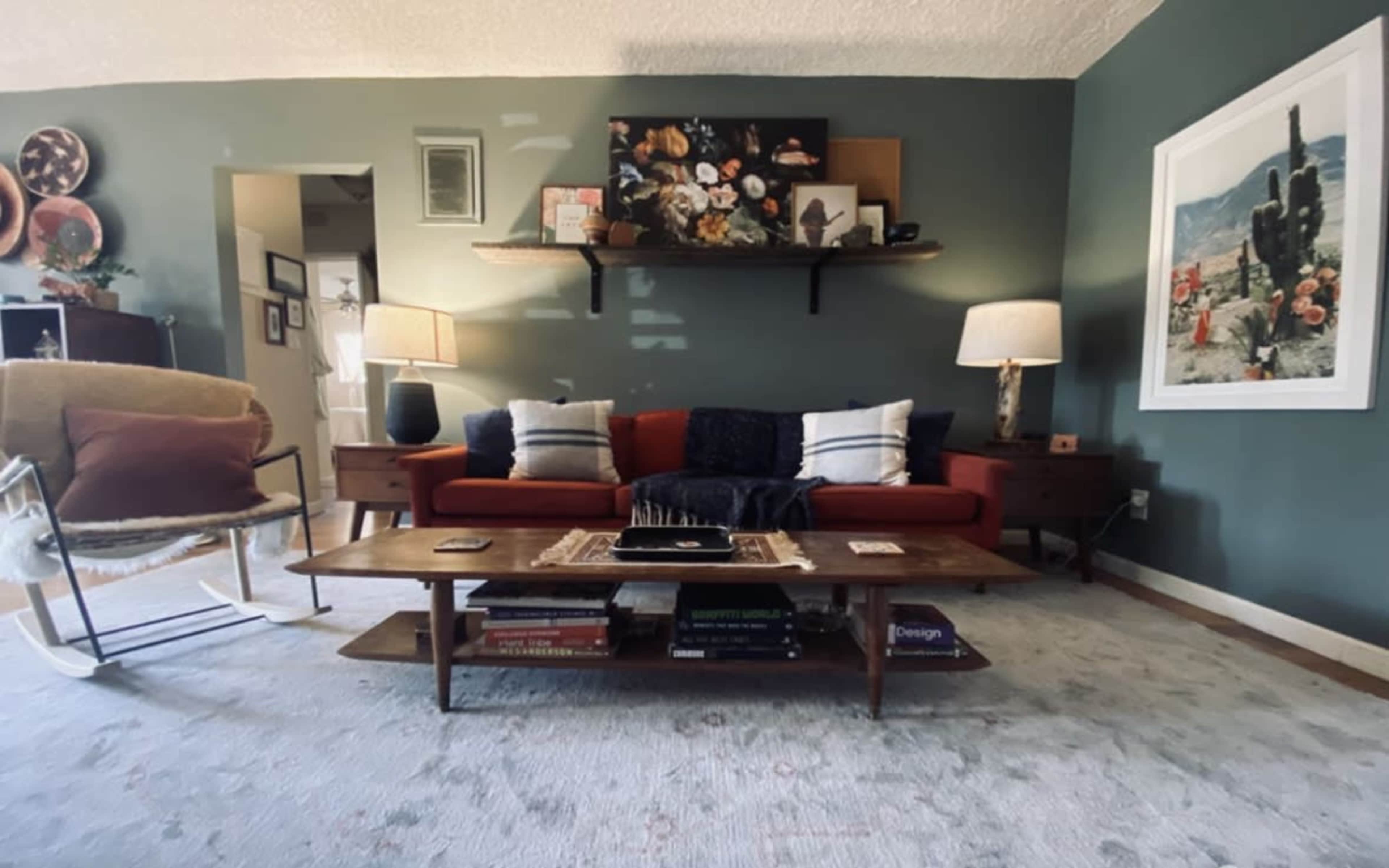 The living room features a red sofa with blue and white throw pillows, a wooden coffee table stacked with books, and two lamps beside the sofa, all against a green wall.