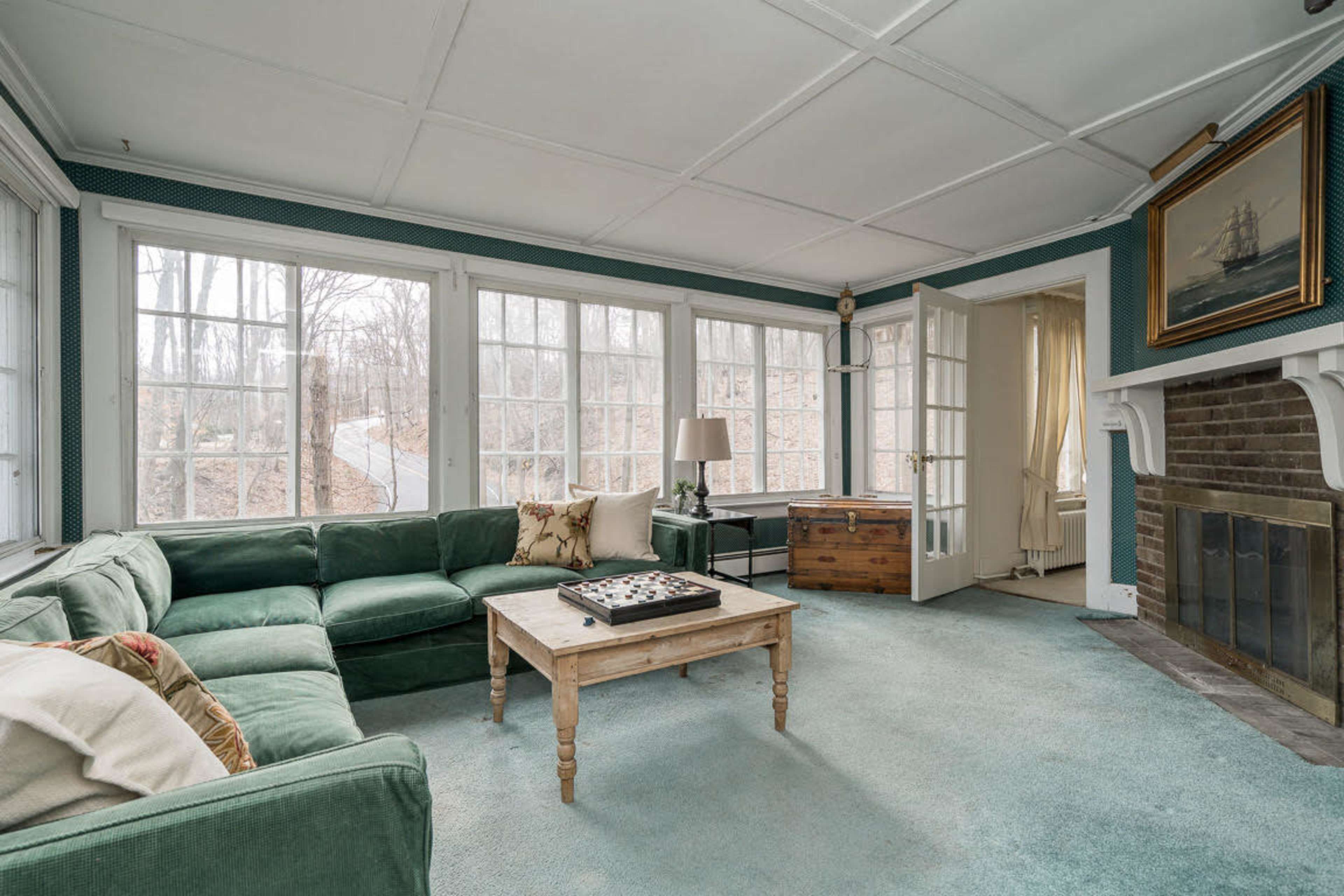 The image shows a sunlit living room with large windows, a green sectional sofa, a coffee table, and a fireplace.
