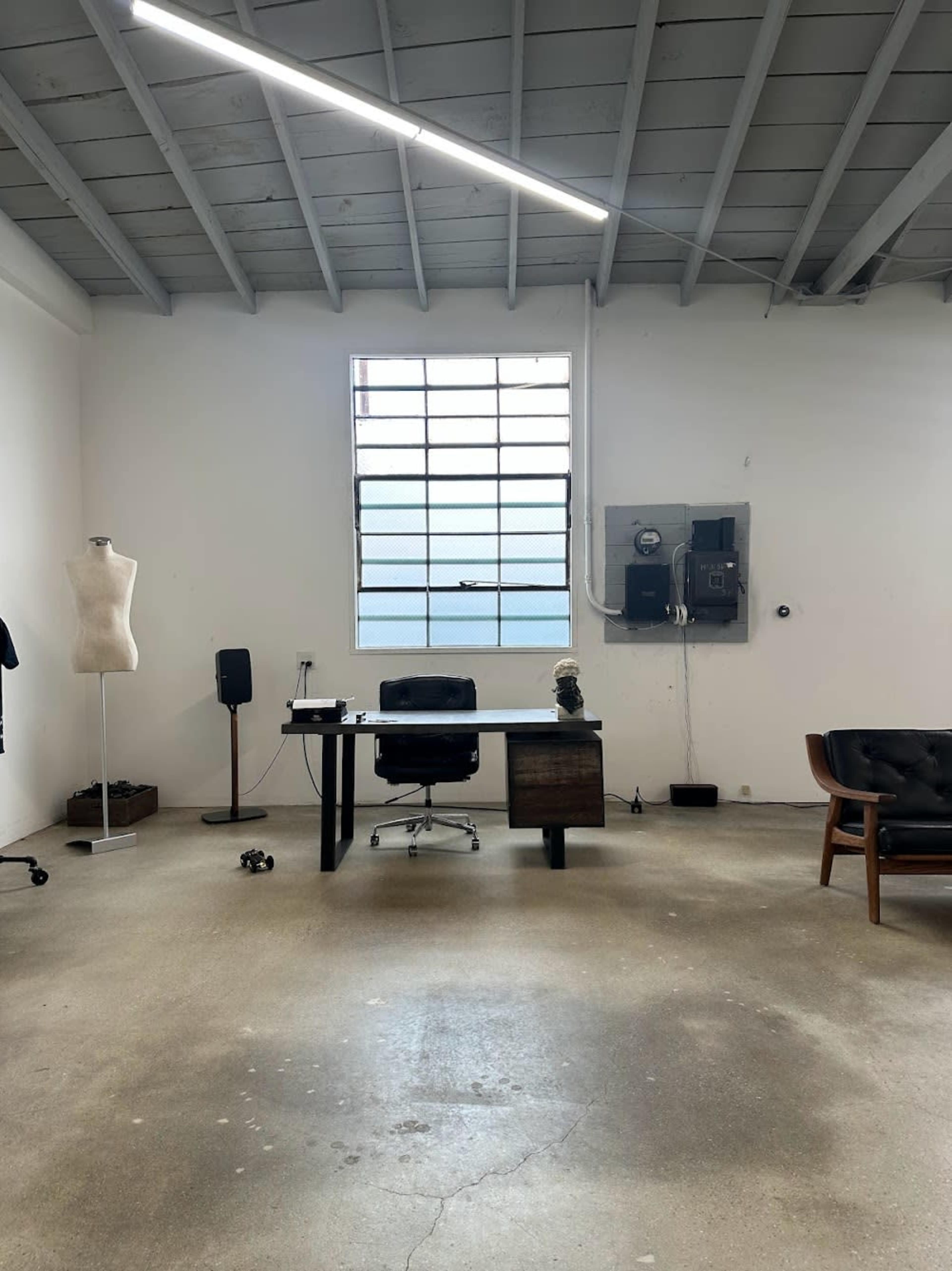 A spacious, minimalistic office with a dark desk and chair, a mannequin, and a large window letting in natural light.