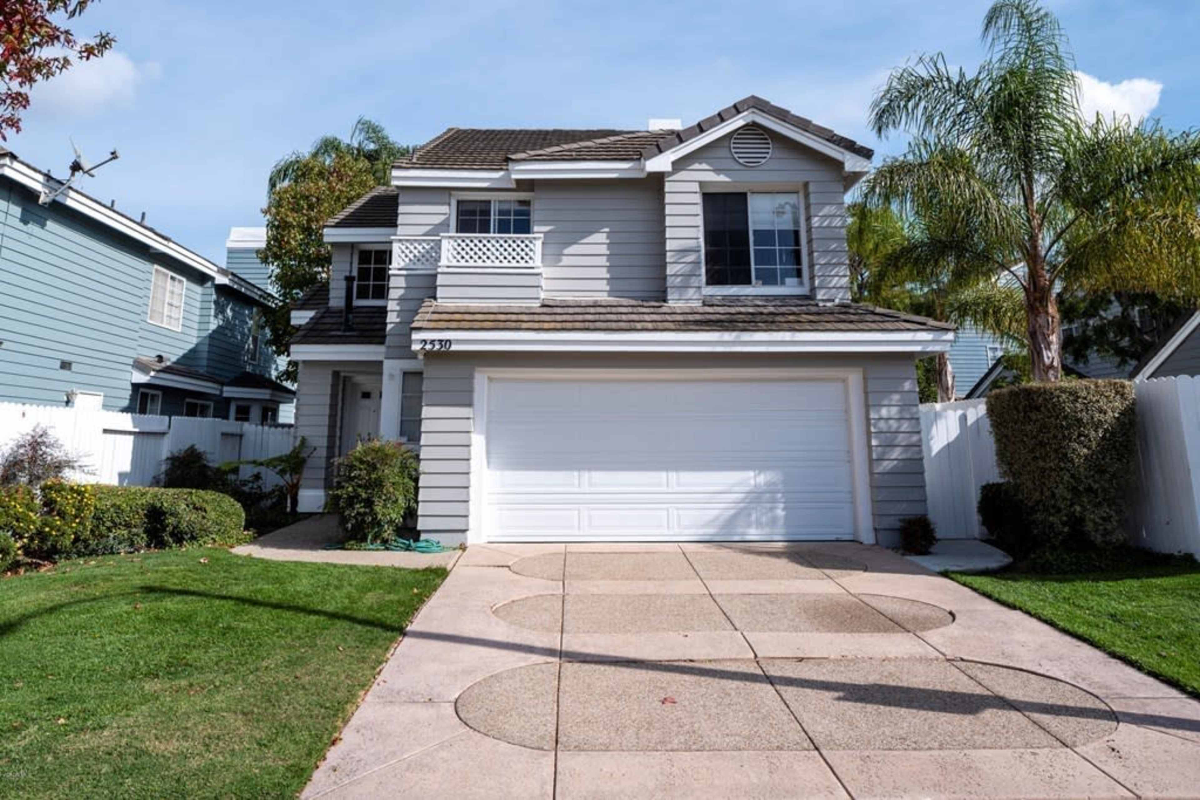 A two-story house with a gray exterior, a white garage door, and palm trees in the front yard.