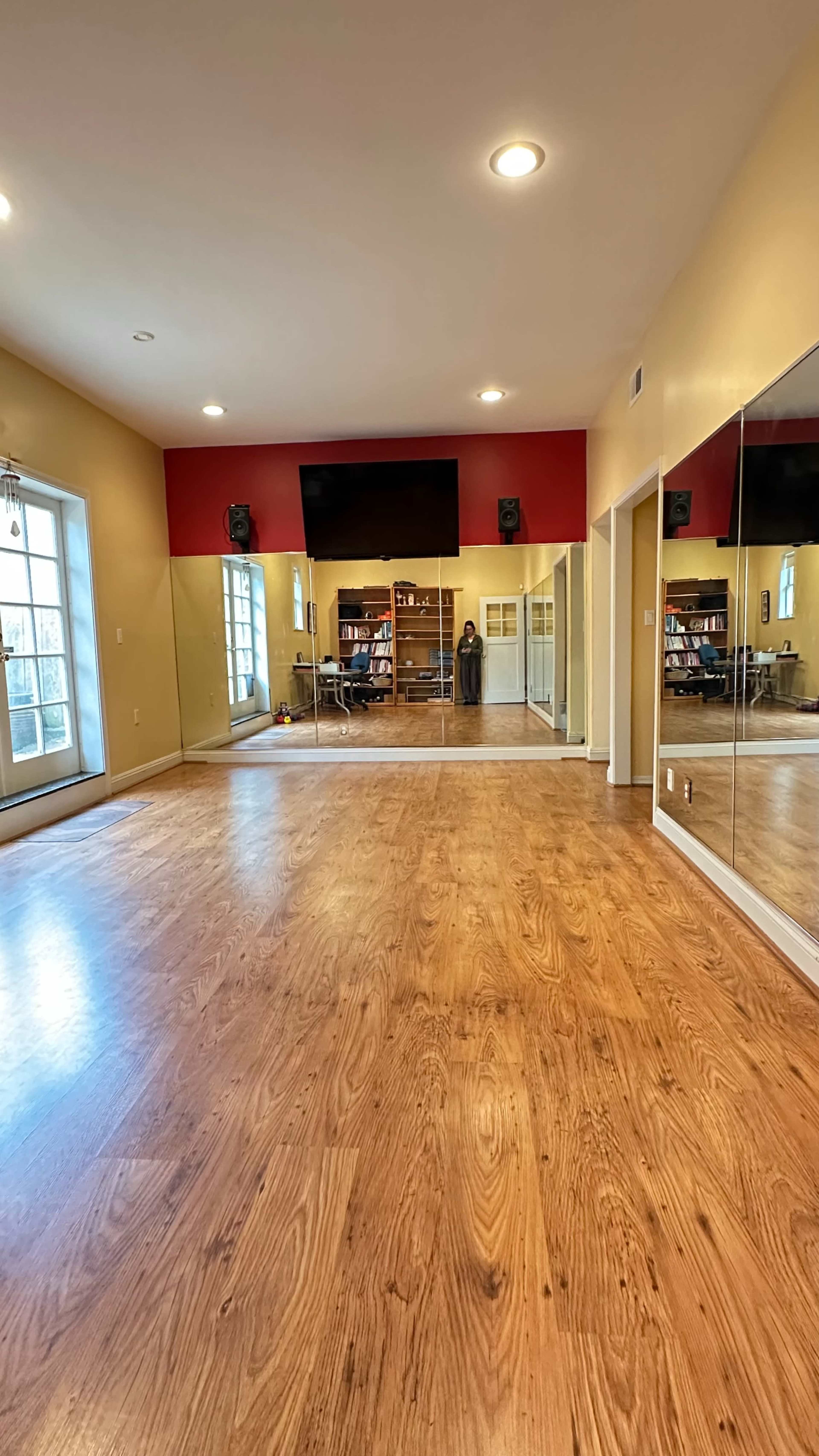The image shows a spacious room with wooden flooring, large mirrors on one wall, and a red accent wall with a television.