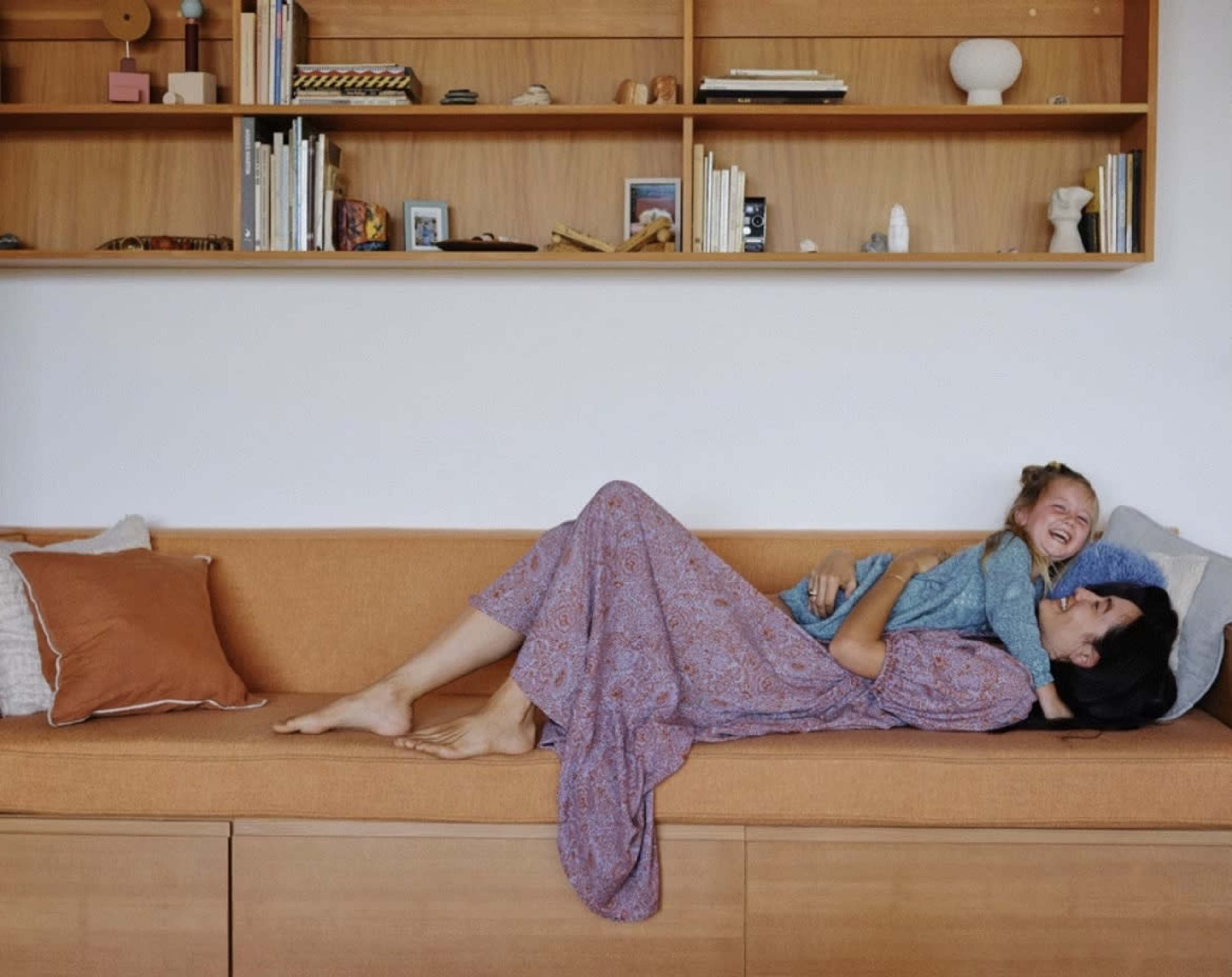 A woman lies on a couch with a young girl laughing beside her, surrounded by decorative items on a shelf above.