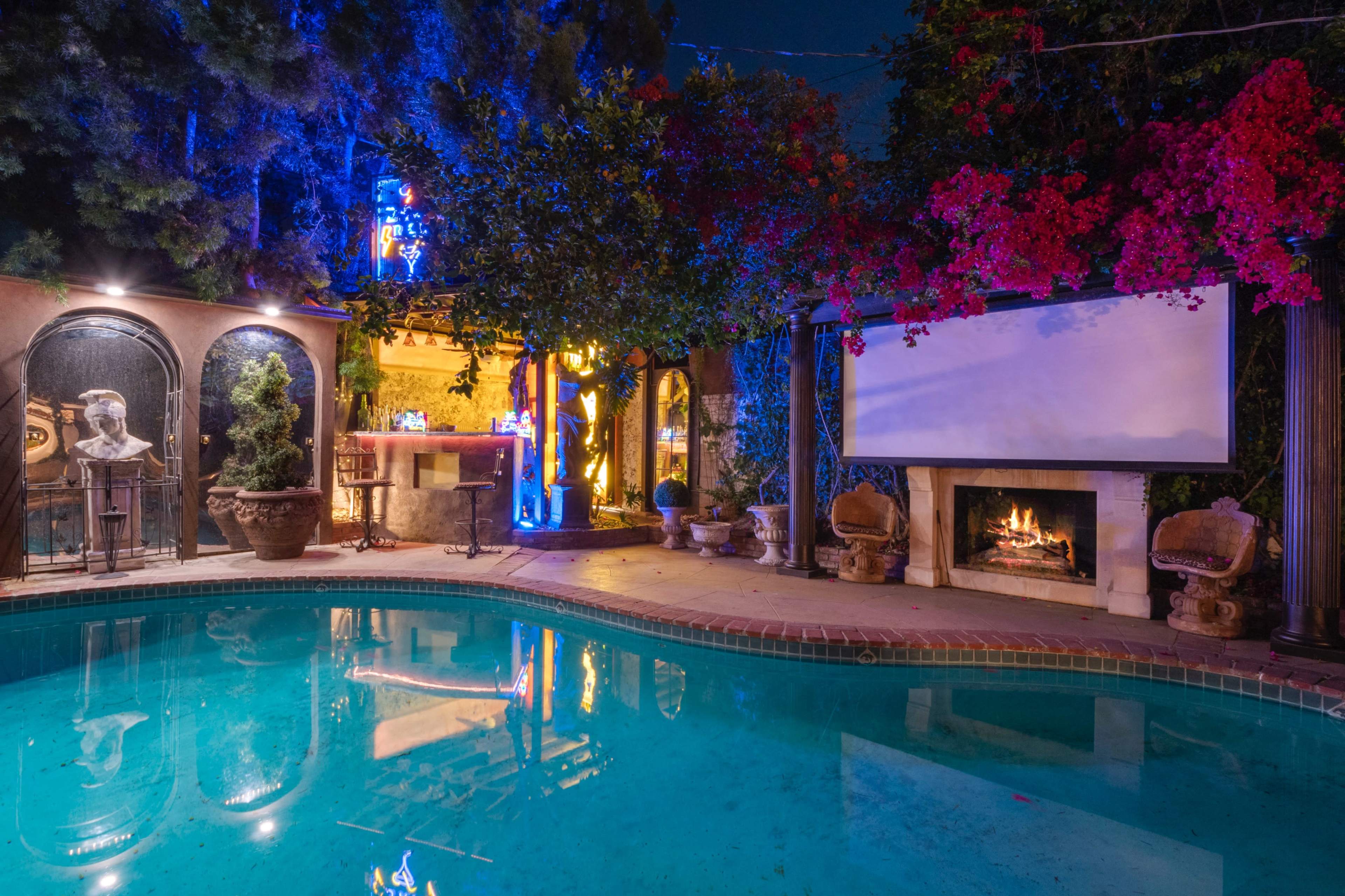 A backyard pool area at night, featuring a swimming pool, a stone fireplace, decorative plants, and a large screen set up near a bar.
