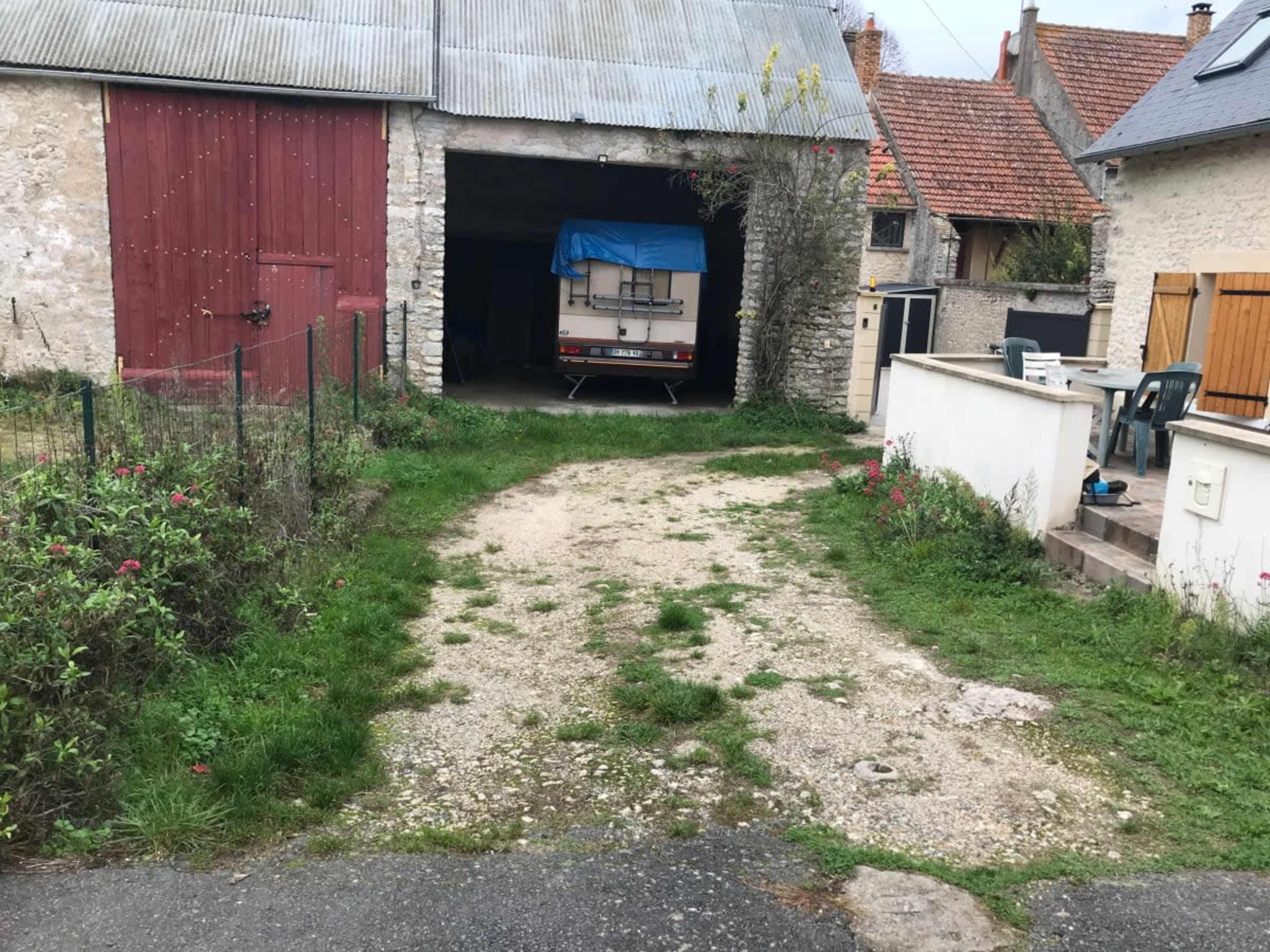 A gravel path leads to a garage with a blue tarpaulin covering a vehicle, surrounded by grass and residential buildings.