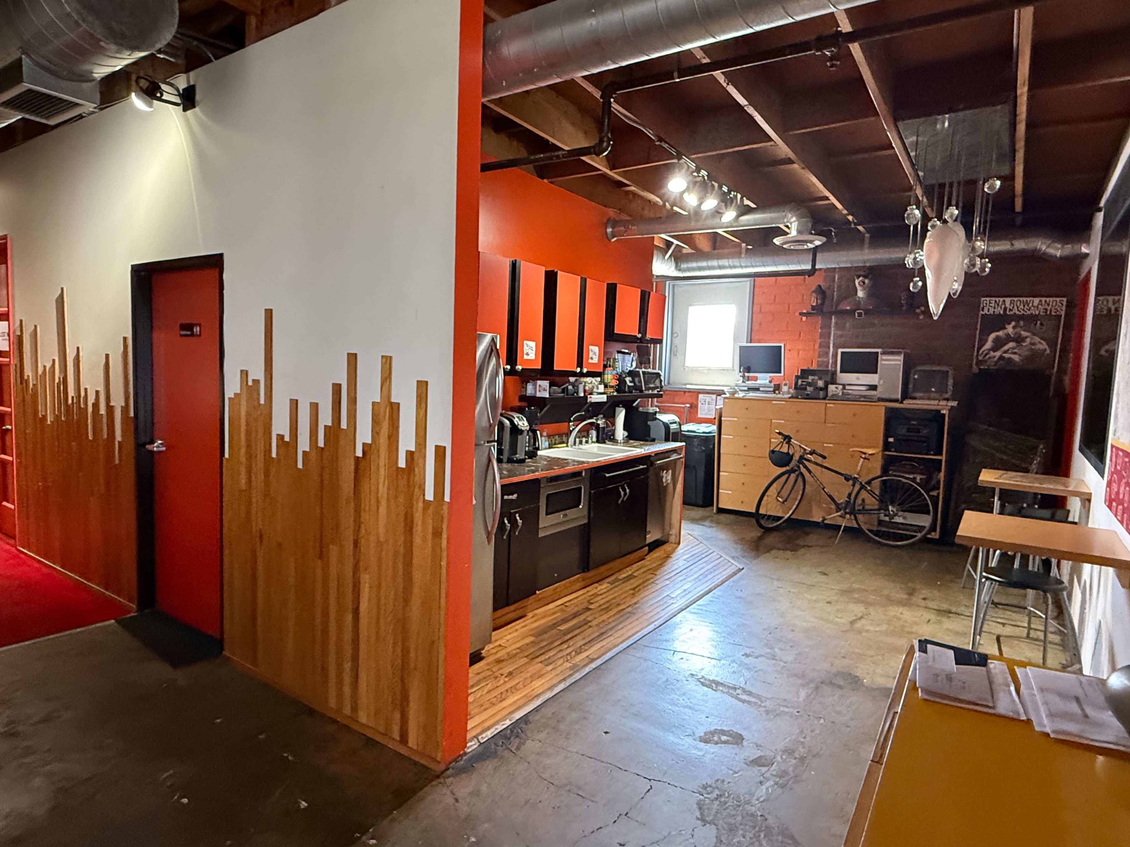 The image shows a spacious kitchen area with red and wood accents, equipped with appliances and a bicycle in the corner, adjacent to a hallway.