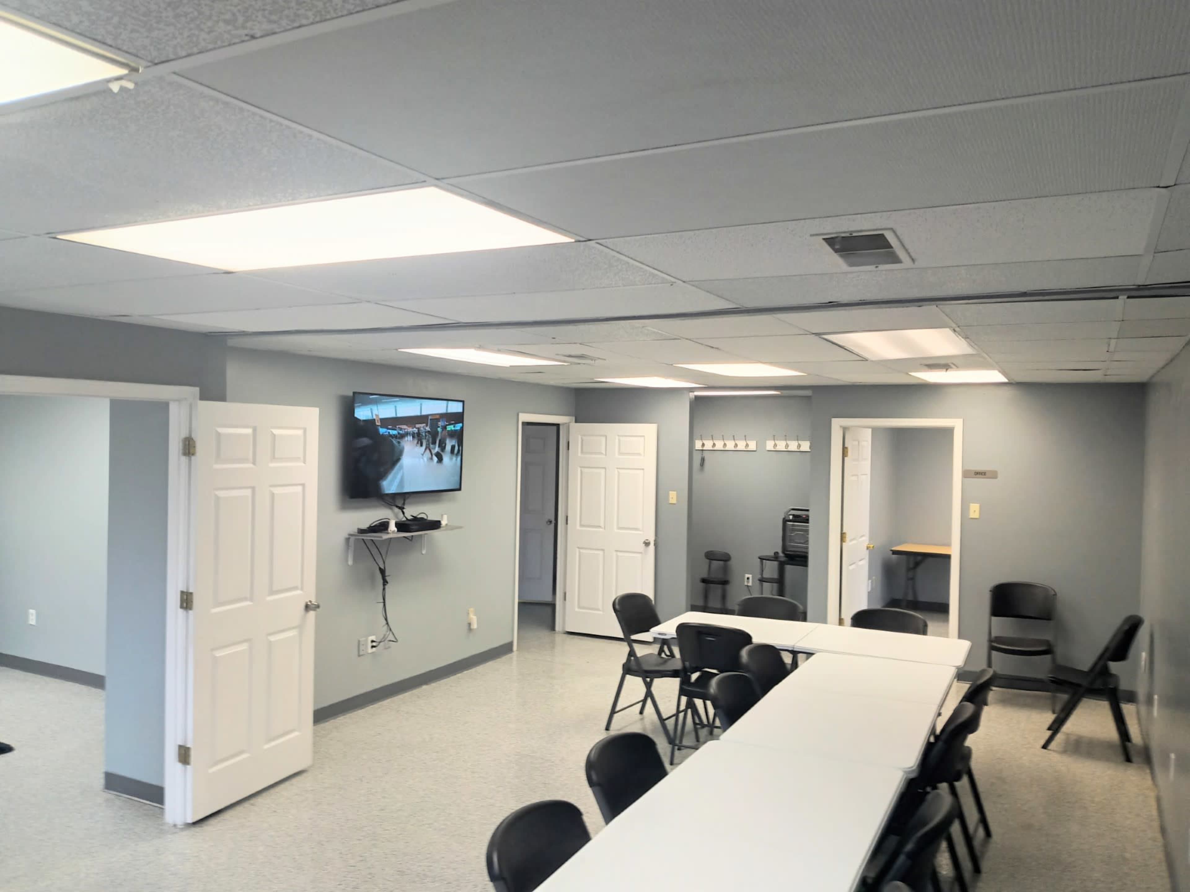 A spacious room with a long white table and black chairs, a TV mounted on the wall, and several doors leading to other areas.