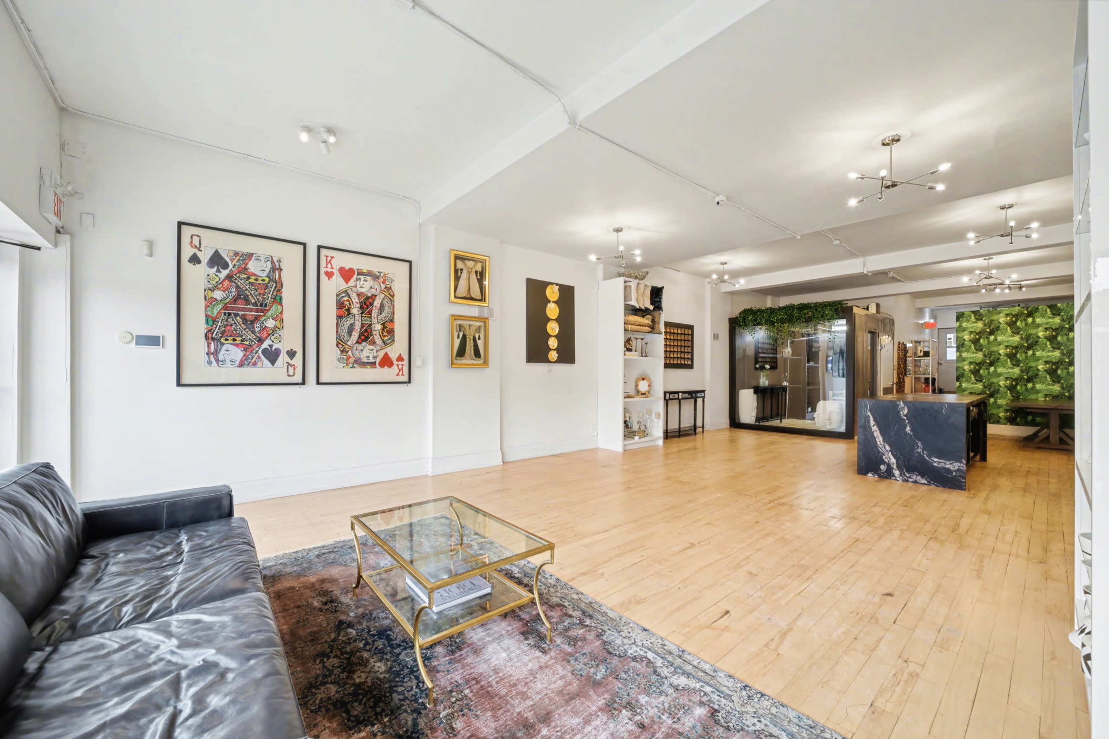 The image depicts a spacious interior with wooden floors, featuring a black leather sofa, artwork on the walls, and an open area that includes a kitchen and dining space.
