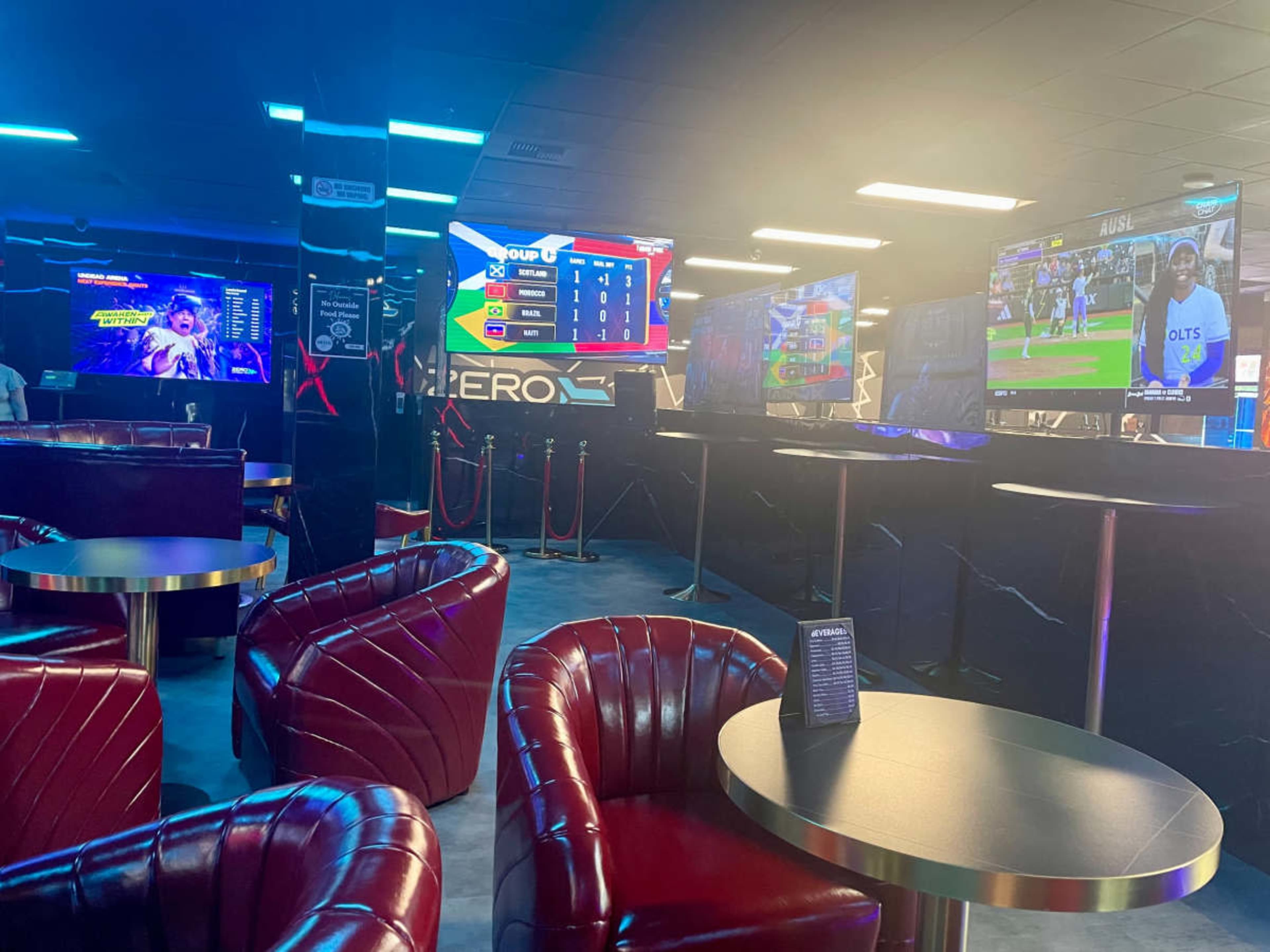The image shows a modern gaming lounge with red leather chairs, tables, and multiple screens displaying video games and sports events.