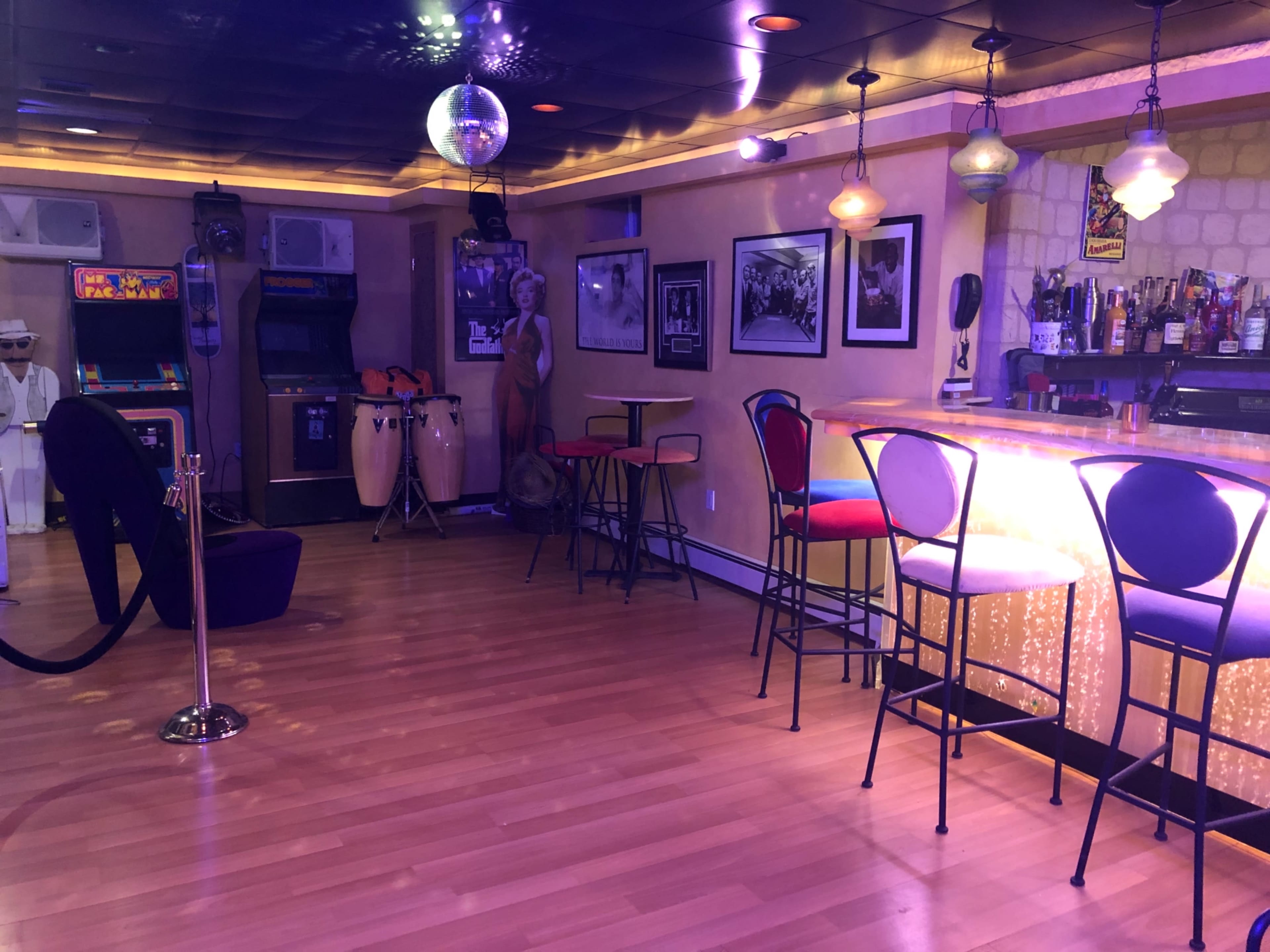 A brightly lit bar area with a seating arrangement of colorful stools, a disco ball hanging from the ceiling, and decorative elements including framed photos and musical instruments.