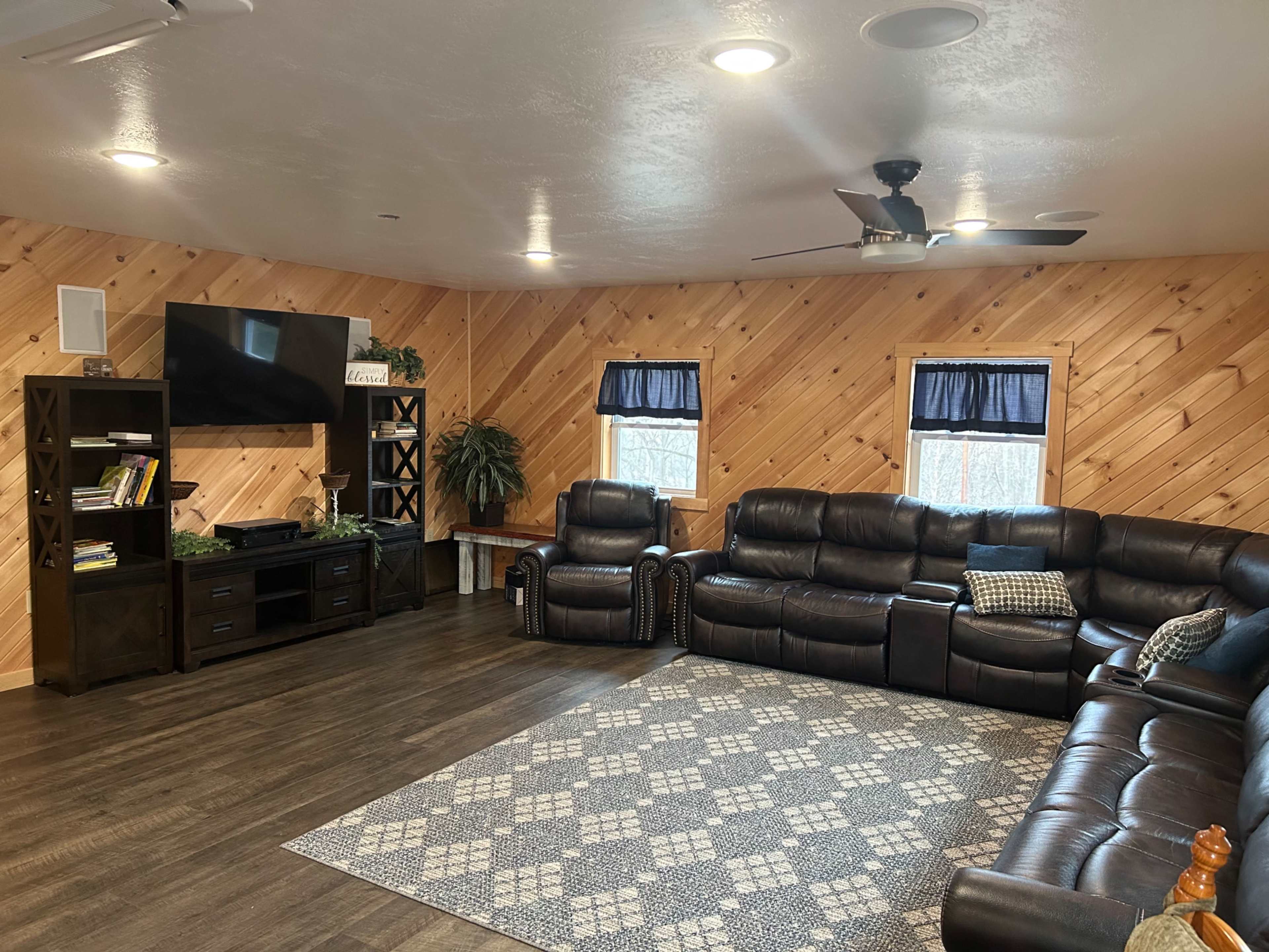 A cozy living room with a dark leather sectional sofa, a black TV stand, and wood-paneled walls, illuminated by recessed lighting.