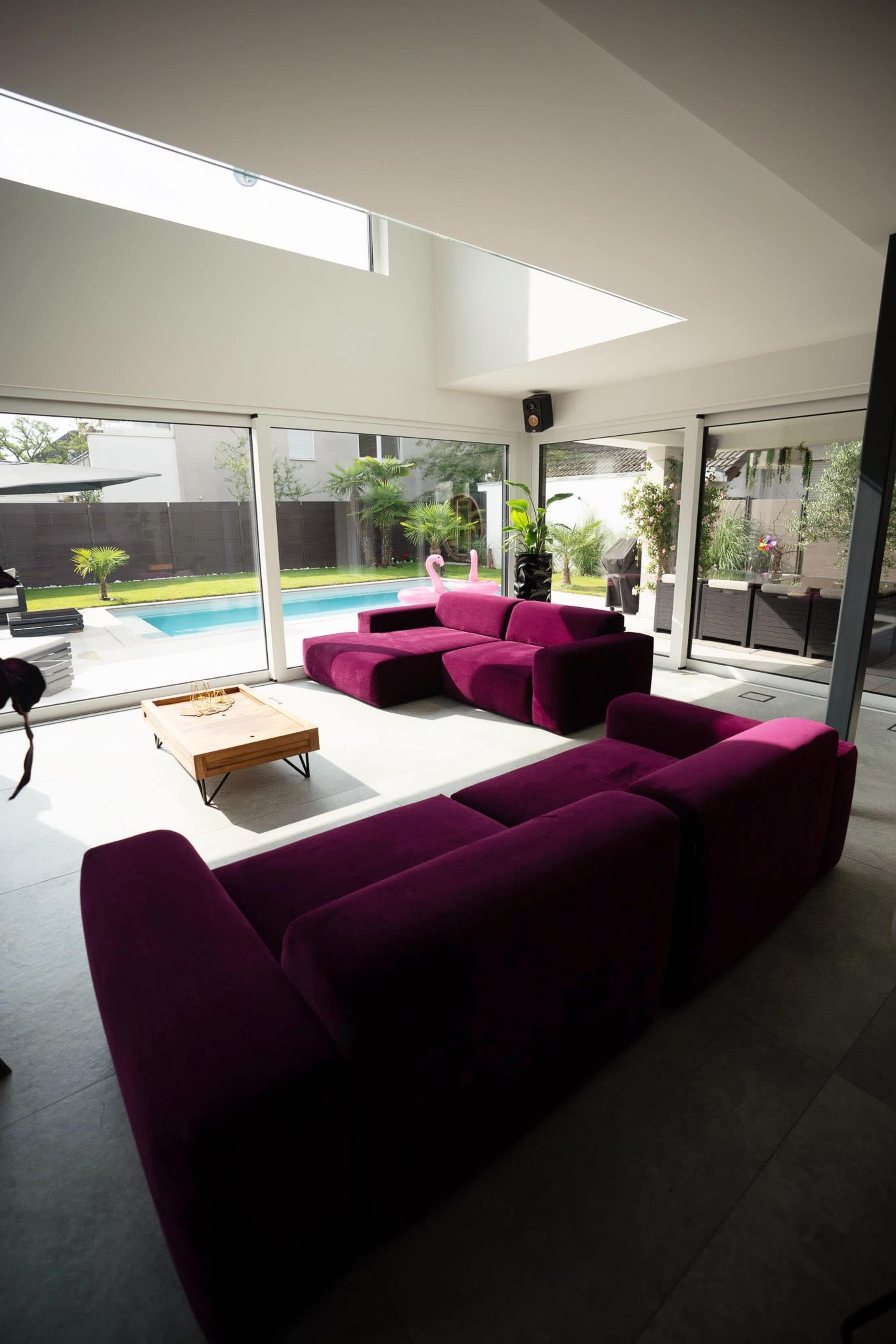 A modern living room features a spacious purple sectional sofa and large windows overlooking a pool area.
