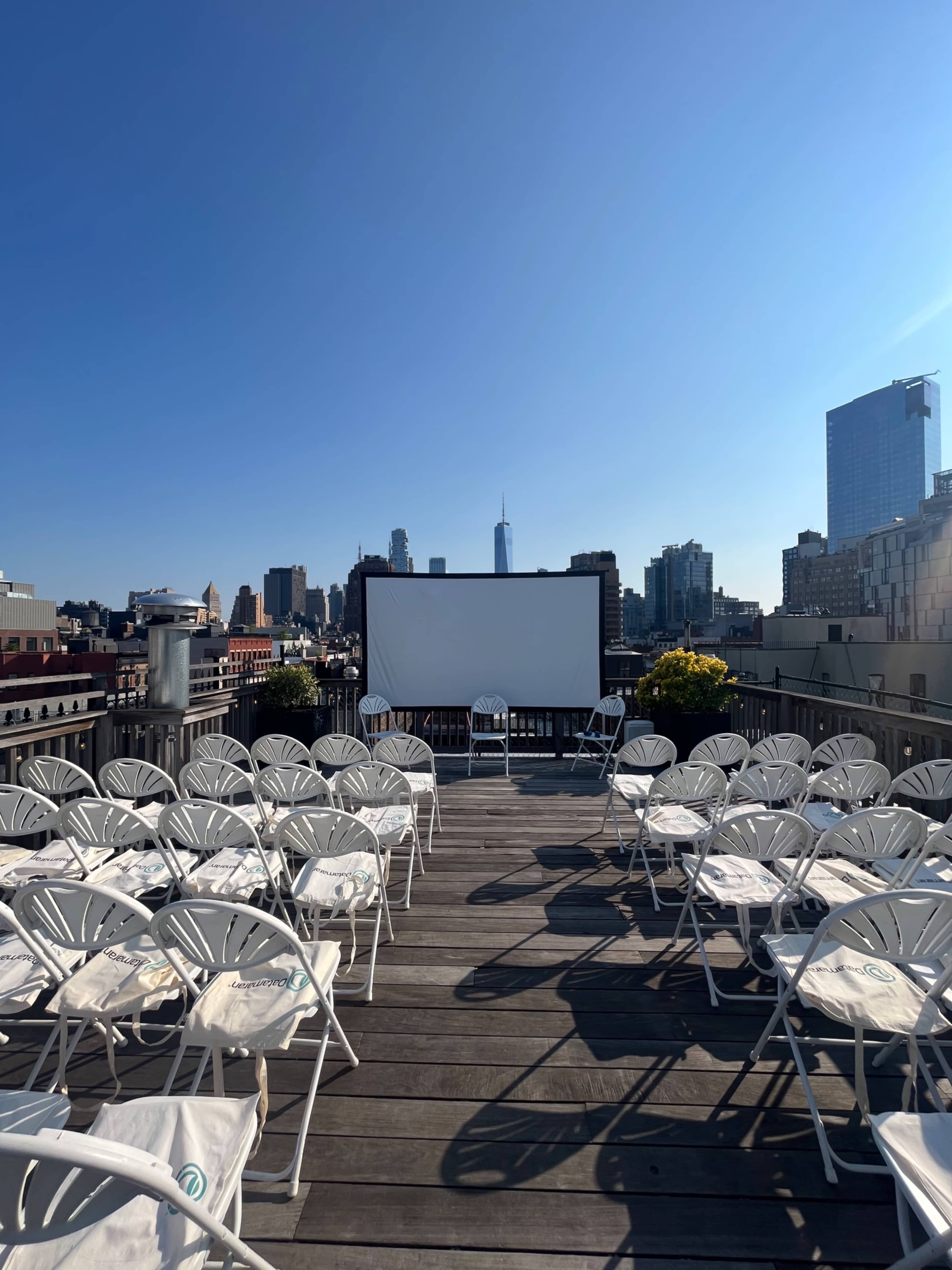 A rooftop cinema setup features rows of white chairs facing a large screen, with a city skyline visible in the background under a clear blue sky.