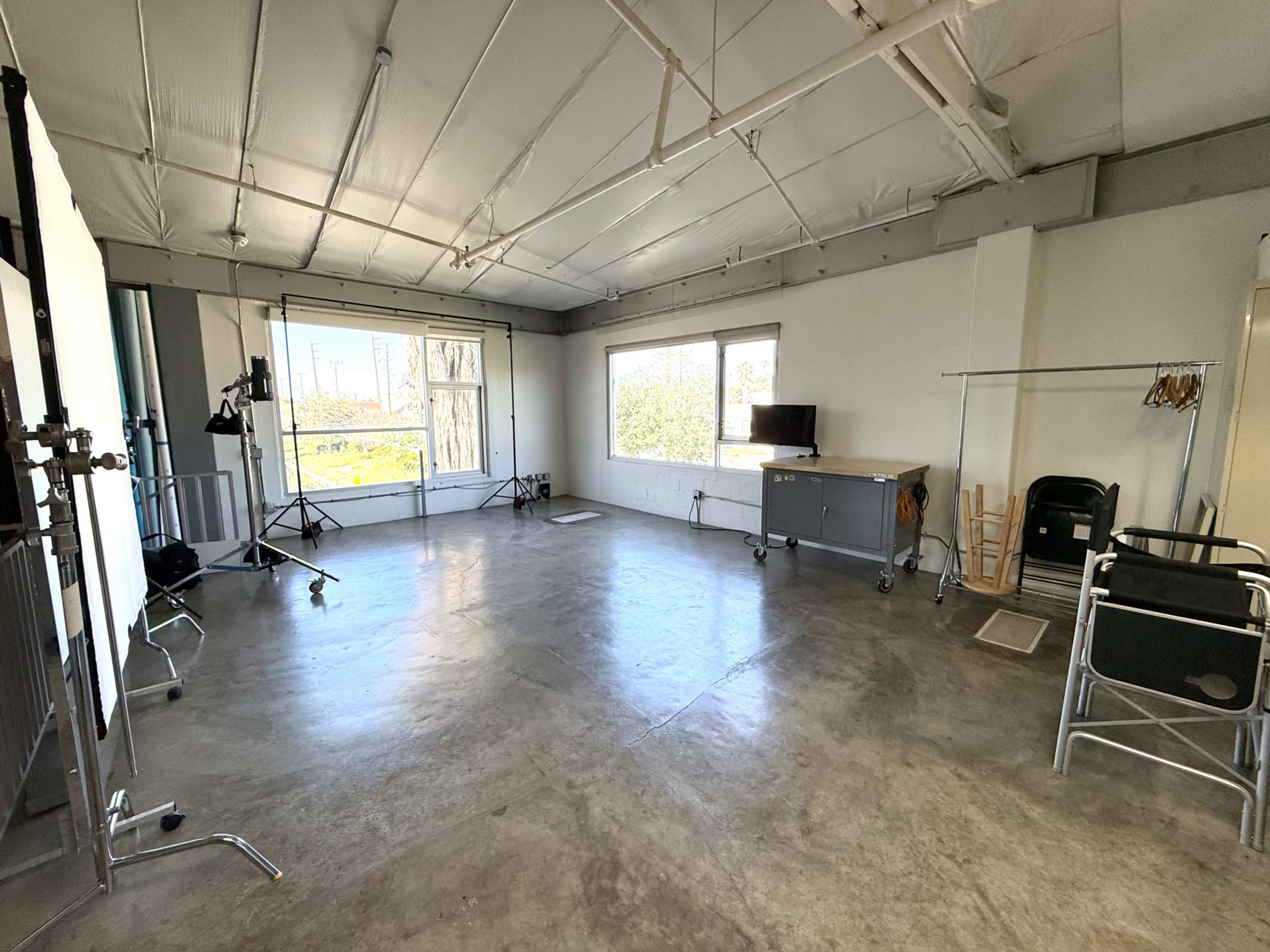 The image shows a spacious photography studio with concrete flooring, large windows, and various equipment including a light stand and a clothing rack.