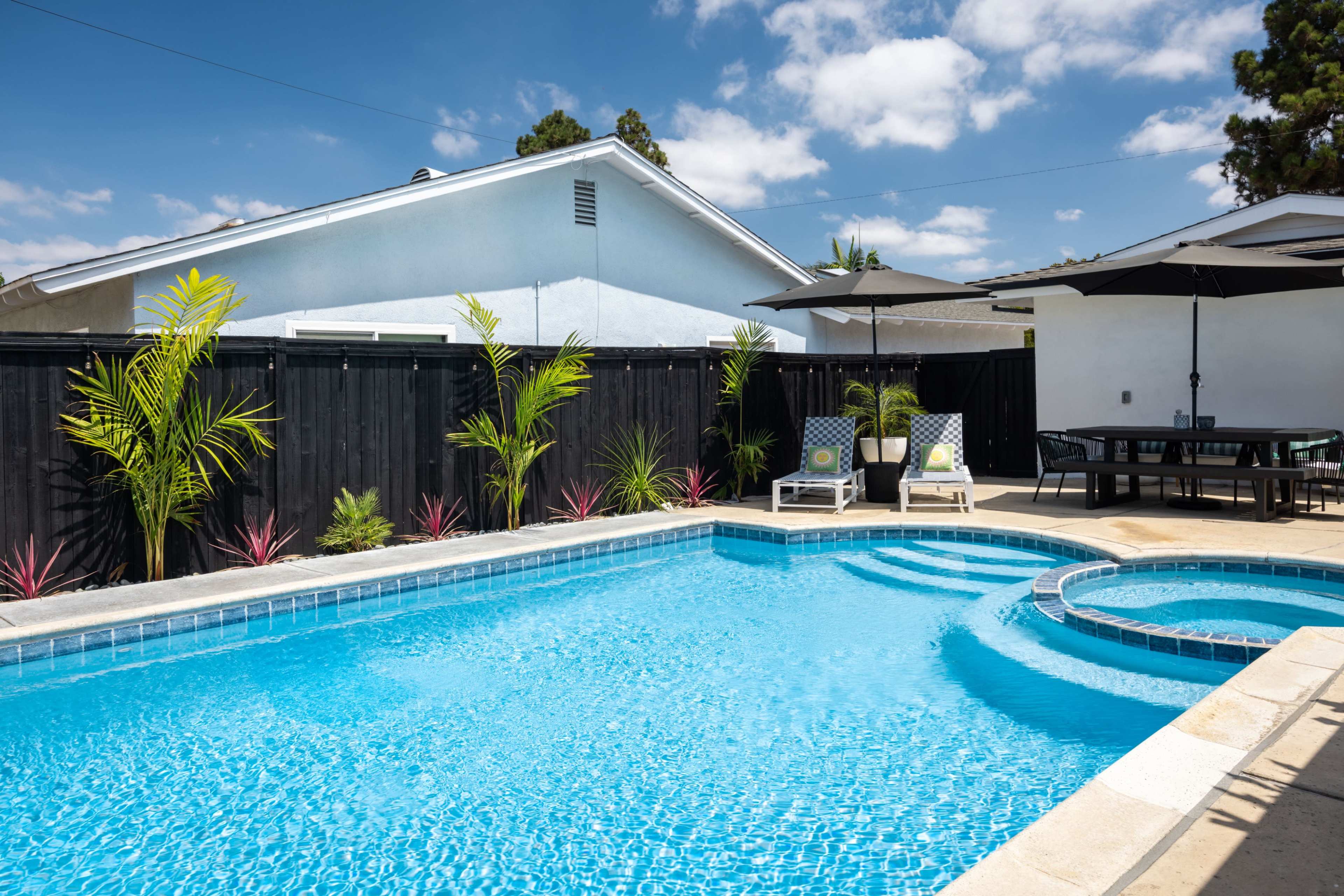 A clear blue swimming pool is surrounded by lounge chairs and tropical plants in a backyard setting.