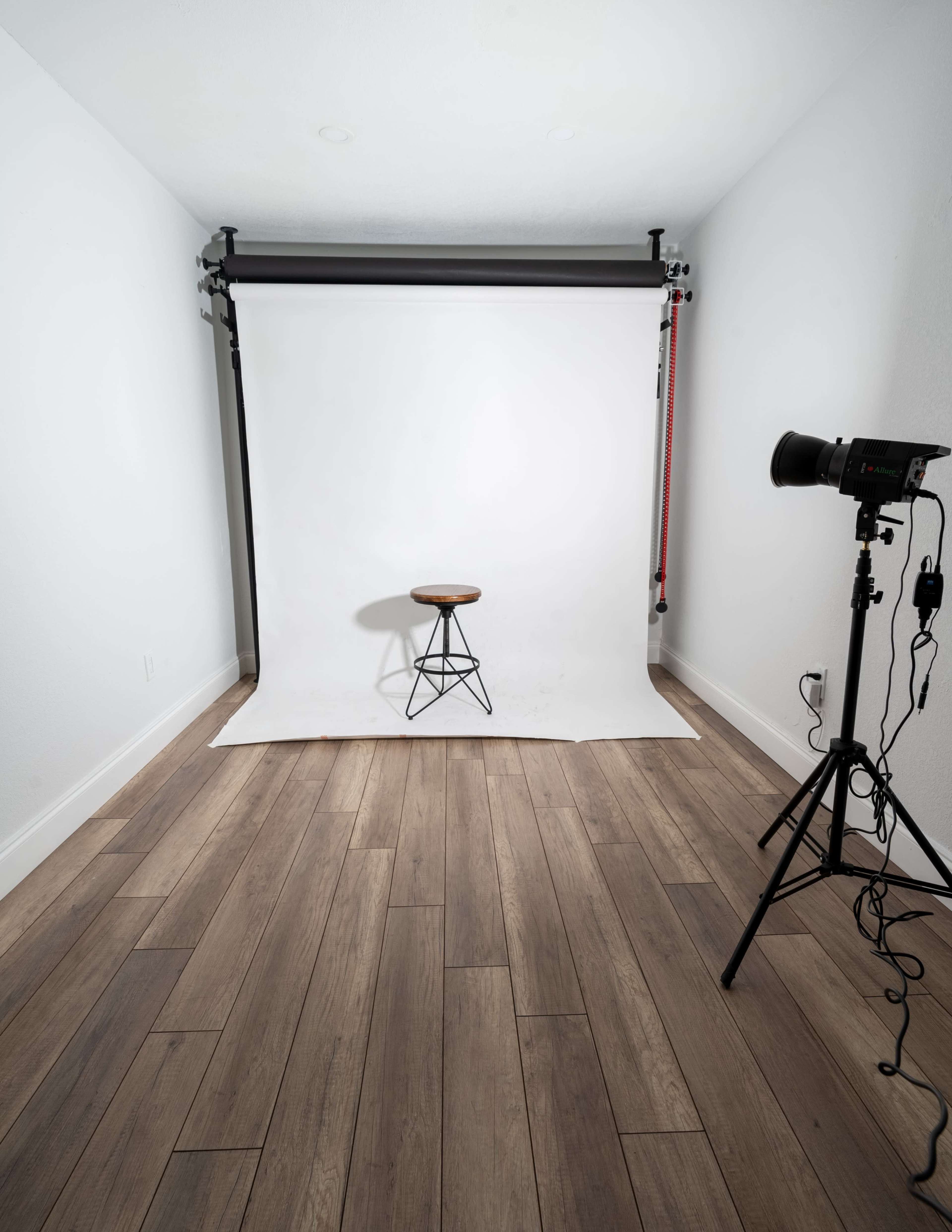 A minimalist studio setup featuring a wooden stool in front of a white backdrop, accompanied by a lighting stand.