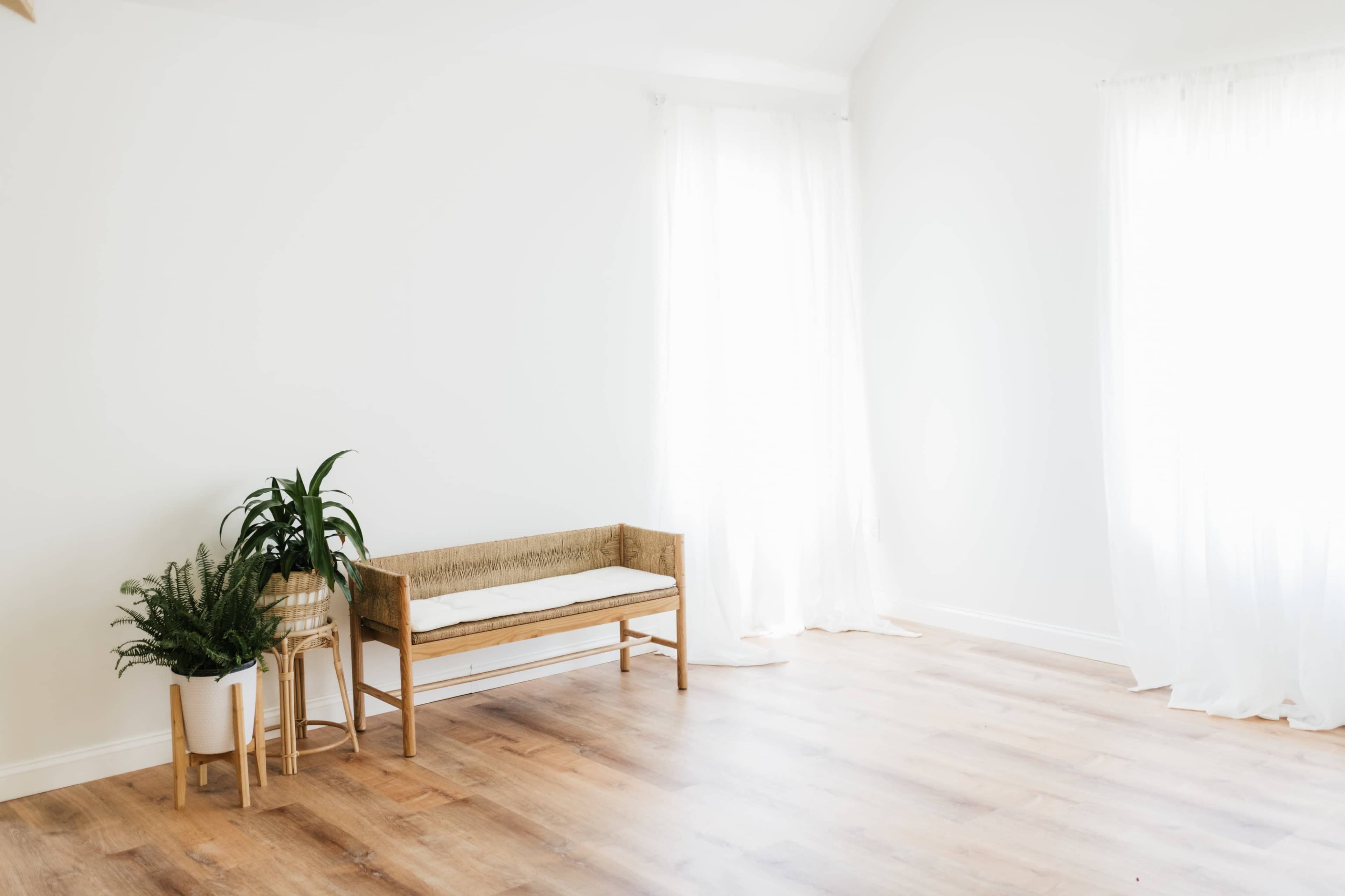 The image shows a minimalist room with a wooden bench, a potted plant, and large windows with sheer curtains allowing natural light to enter.