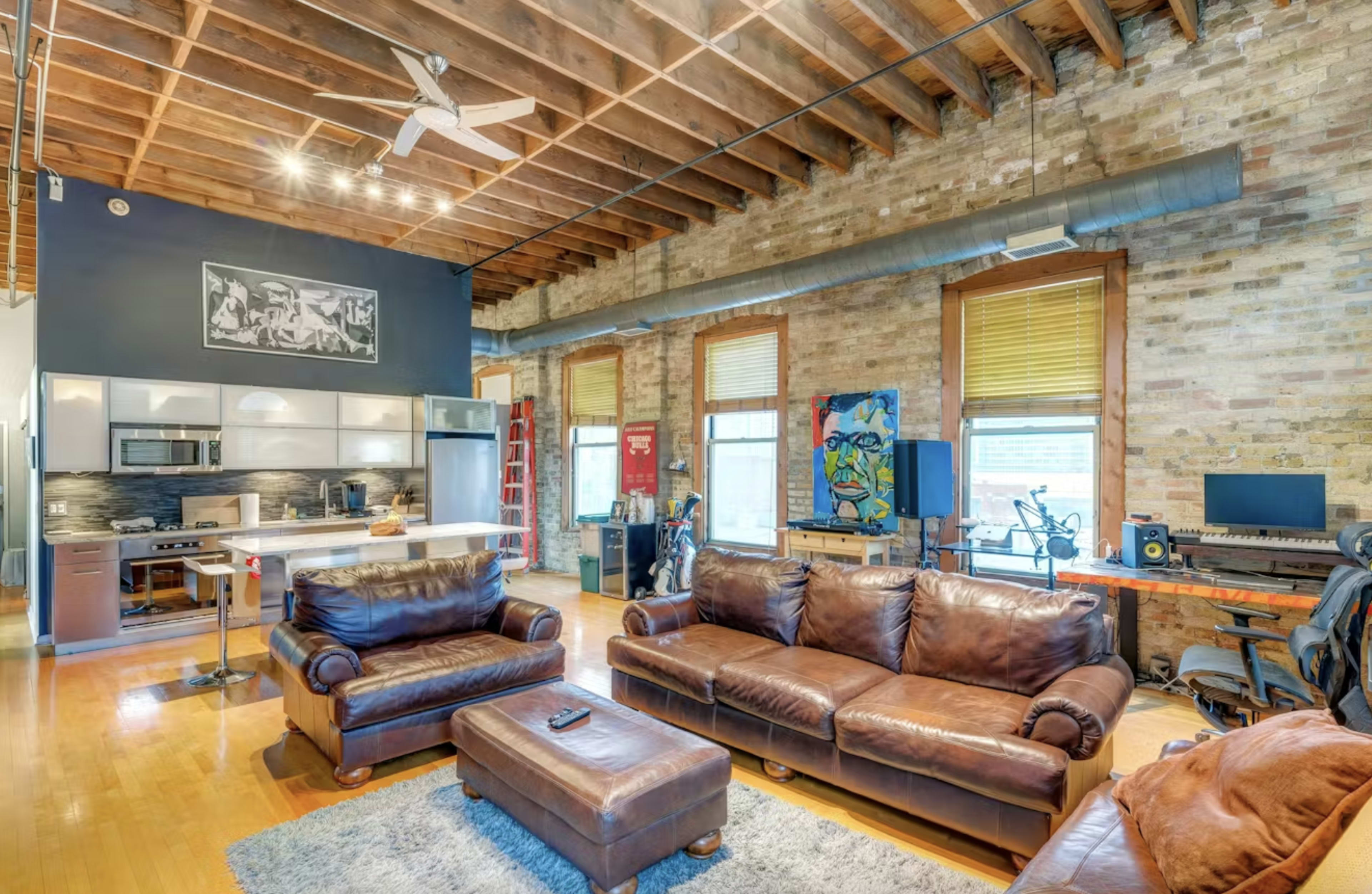 The image shows a spacious living area featuring leather sofas, a coffee table, and an open kitchen with modern appliances, all set against a backdrop of exposed brick walls and wooden beams.