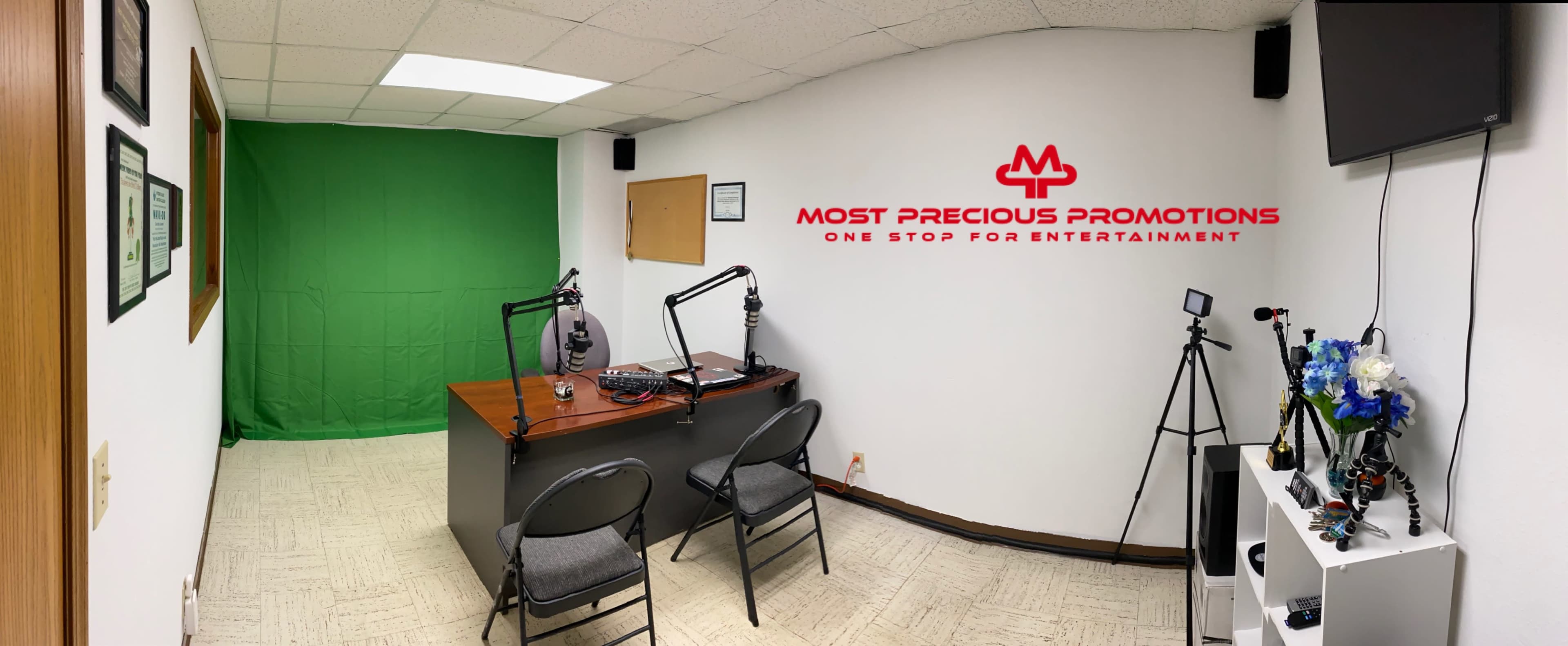 A small recording studio features a green screen, two chairs, microphones on stands, a camera, and a decorative arrangement of flowers.