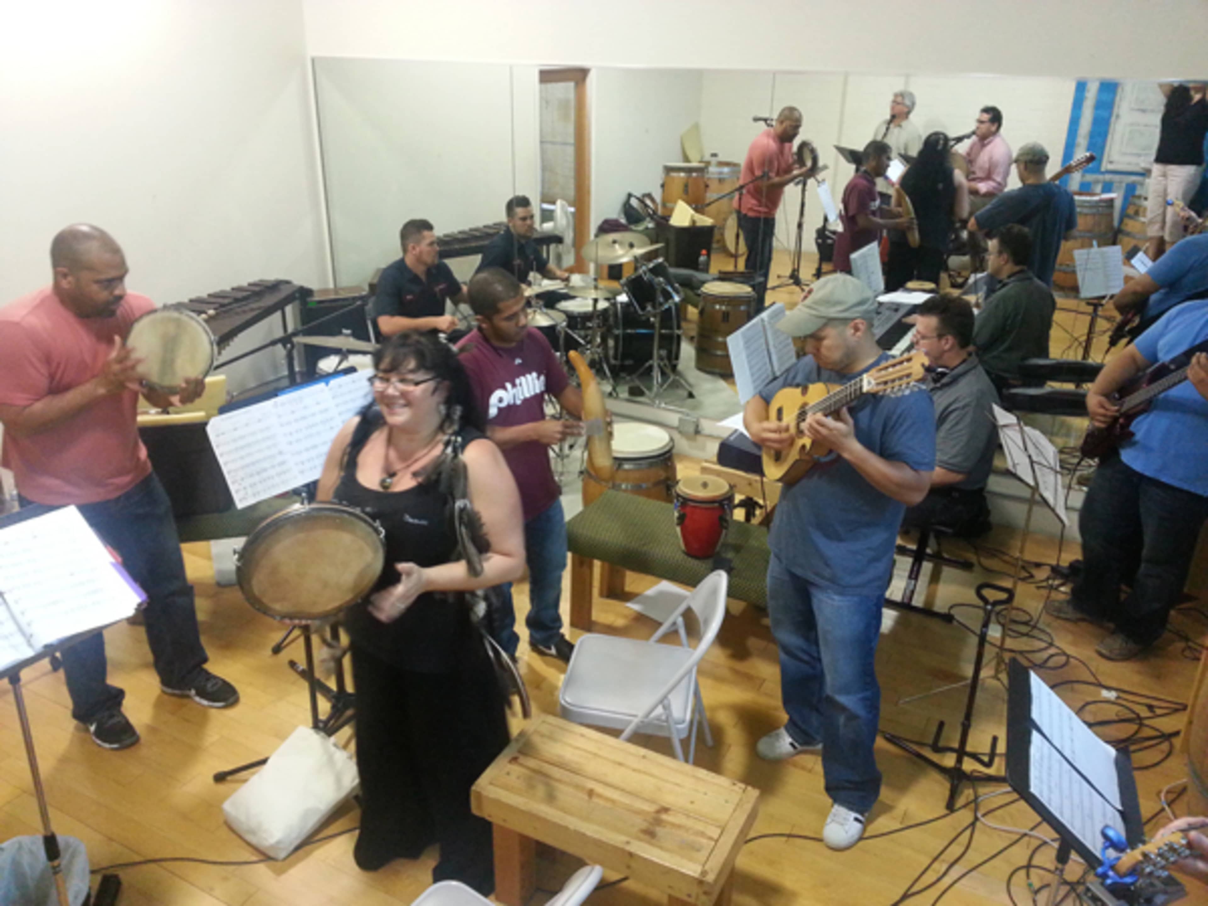 A group of musicians plays various instruments in a rehearsal space filled with chairs and music stands.