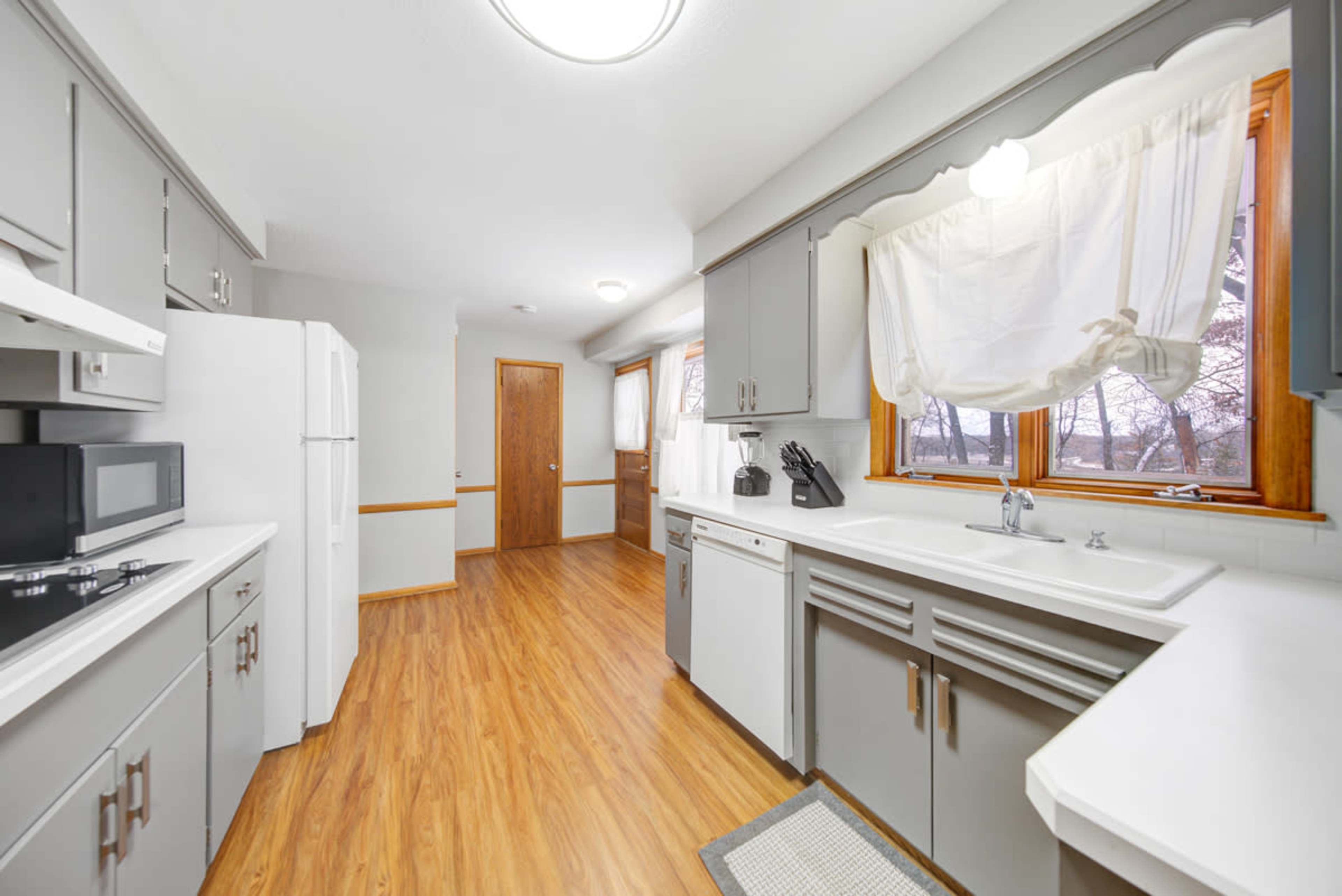 The kitchen features grey cabinetry, a white sink, and a window that provides natural light, with a door leading to another room in the background.