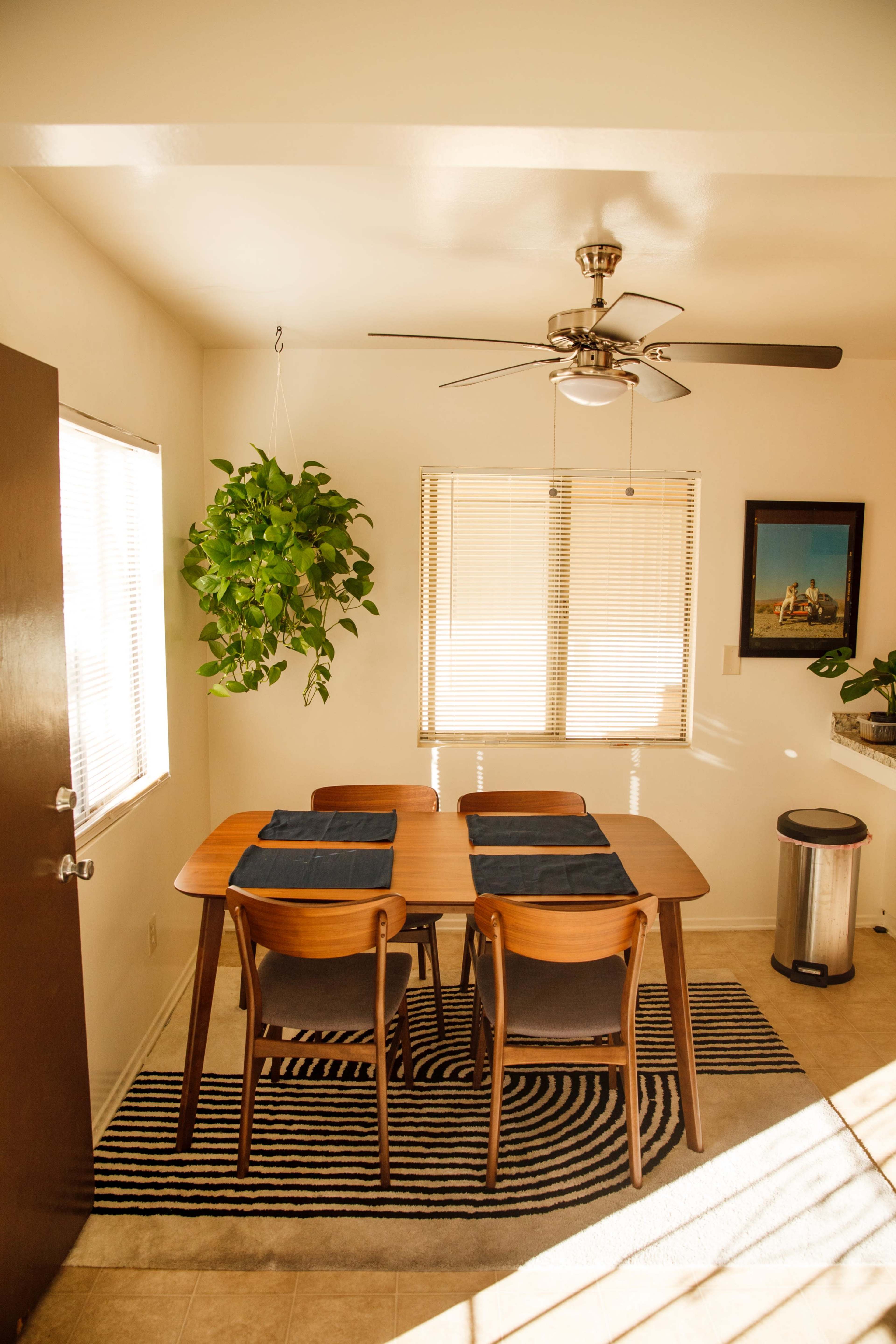 A dining area features a wooden table with four chairs, a ceiling fan, a potted plant hanging near a window, and a trash can beside the table.