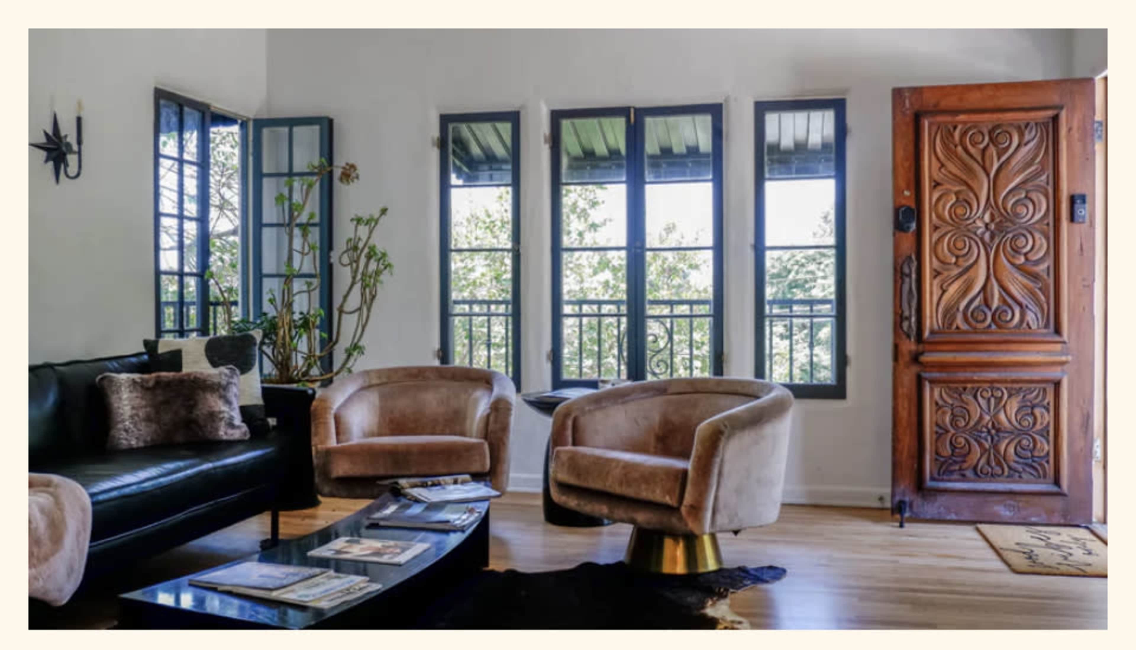 The image shows a modern living room featuring two comfortable armchairs, a sleek coffee table with magazines, and a large wooden door with intricate carvings, all surrounded by bright windows.