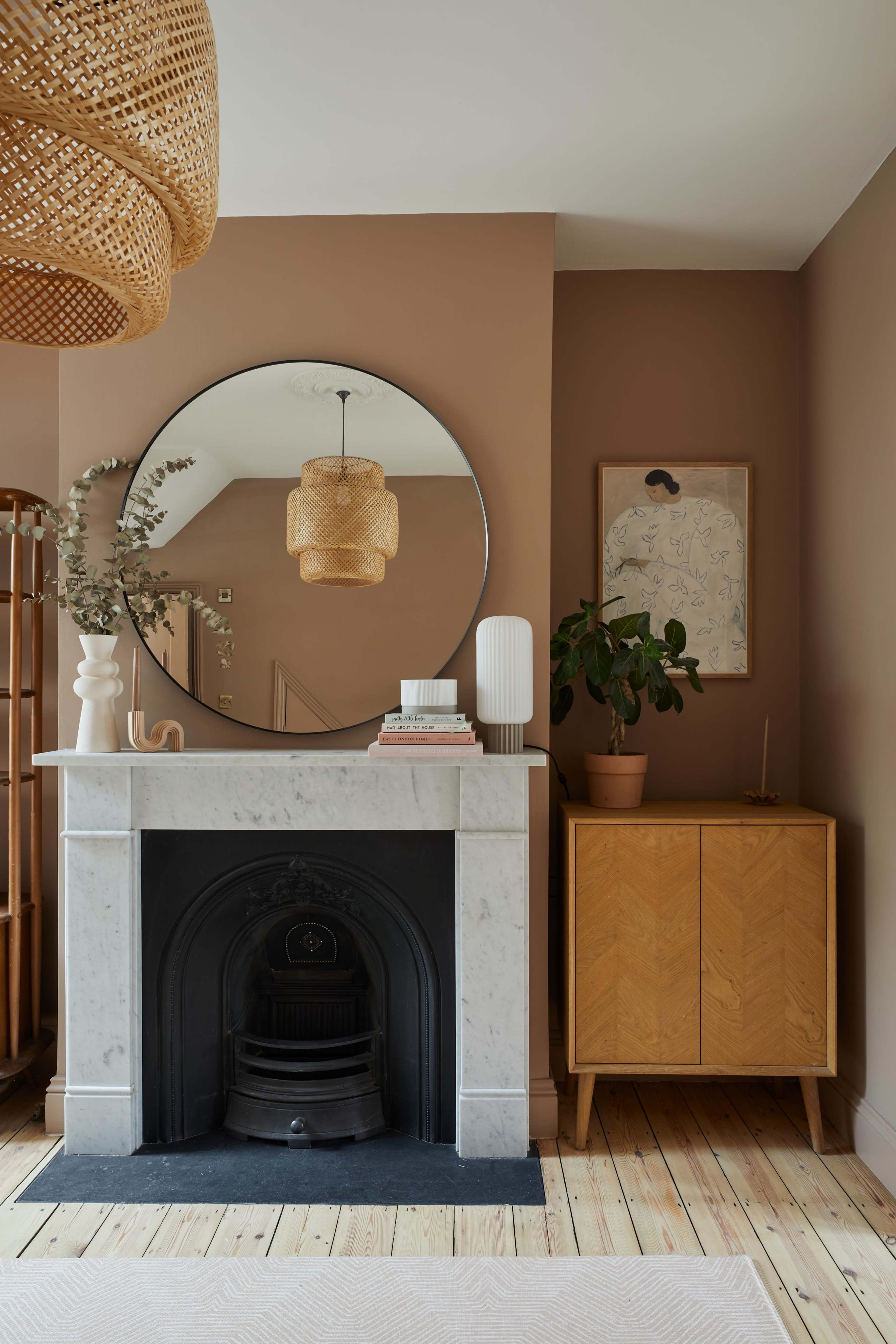 A living room with a round mirror above a marble fireplace, a woven pendant light, and a wooden cabinet alongside a potted plant.