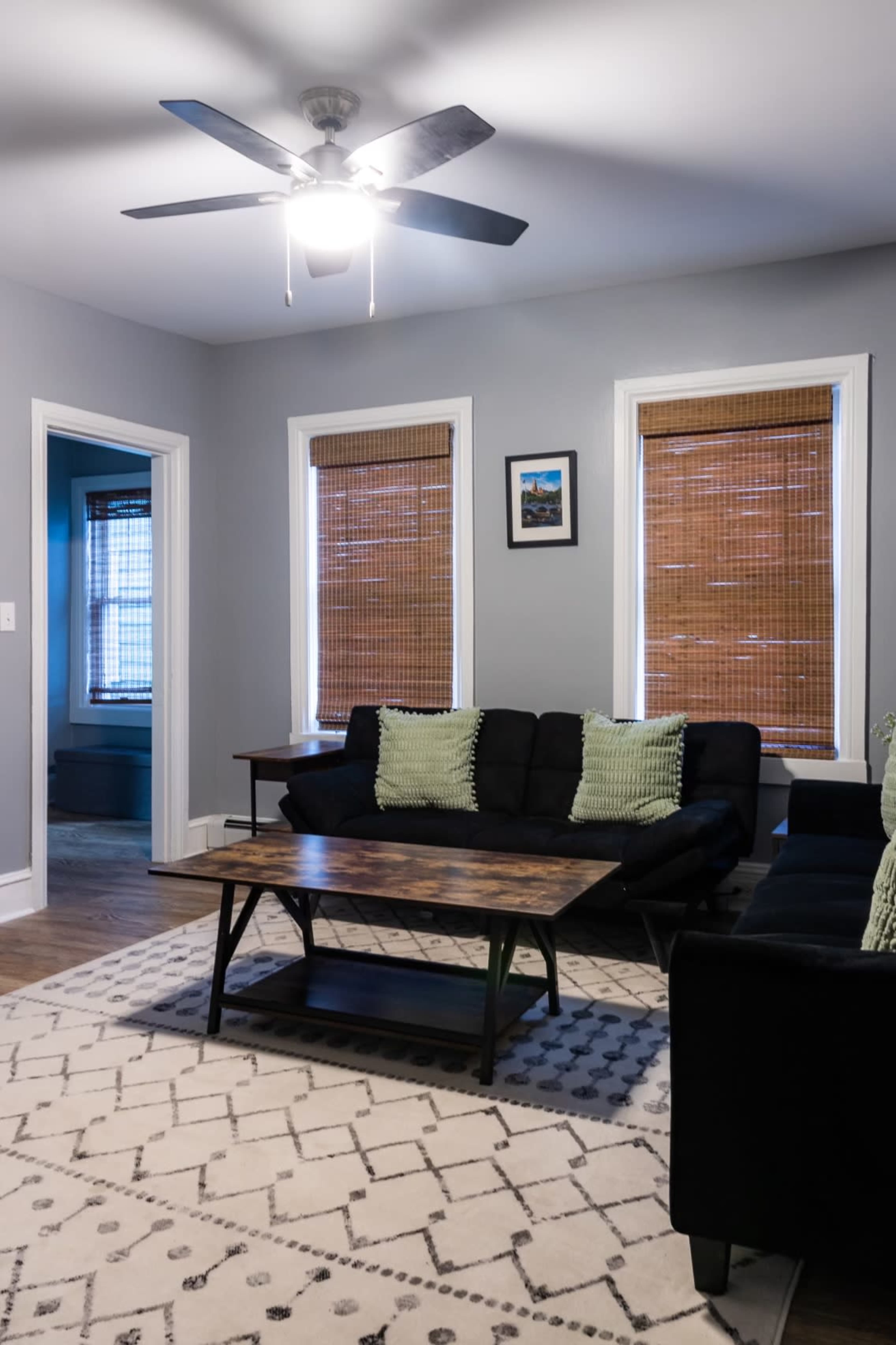 A living room features a black couch with green cushions, a wooden coffee table, and natural light coming through window blinds.