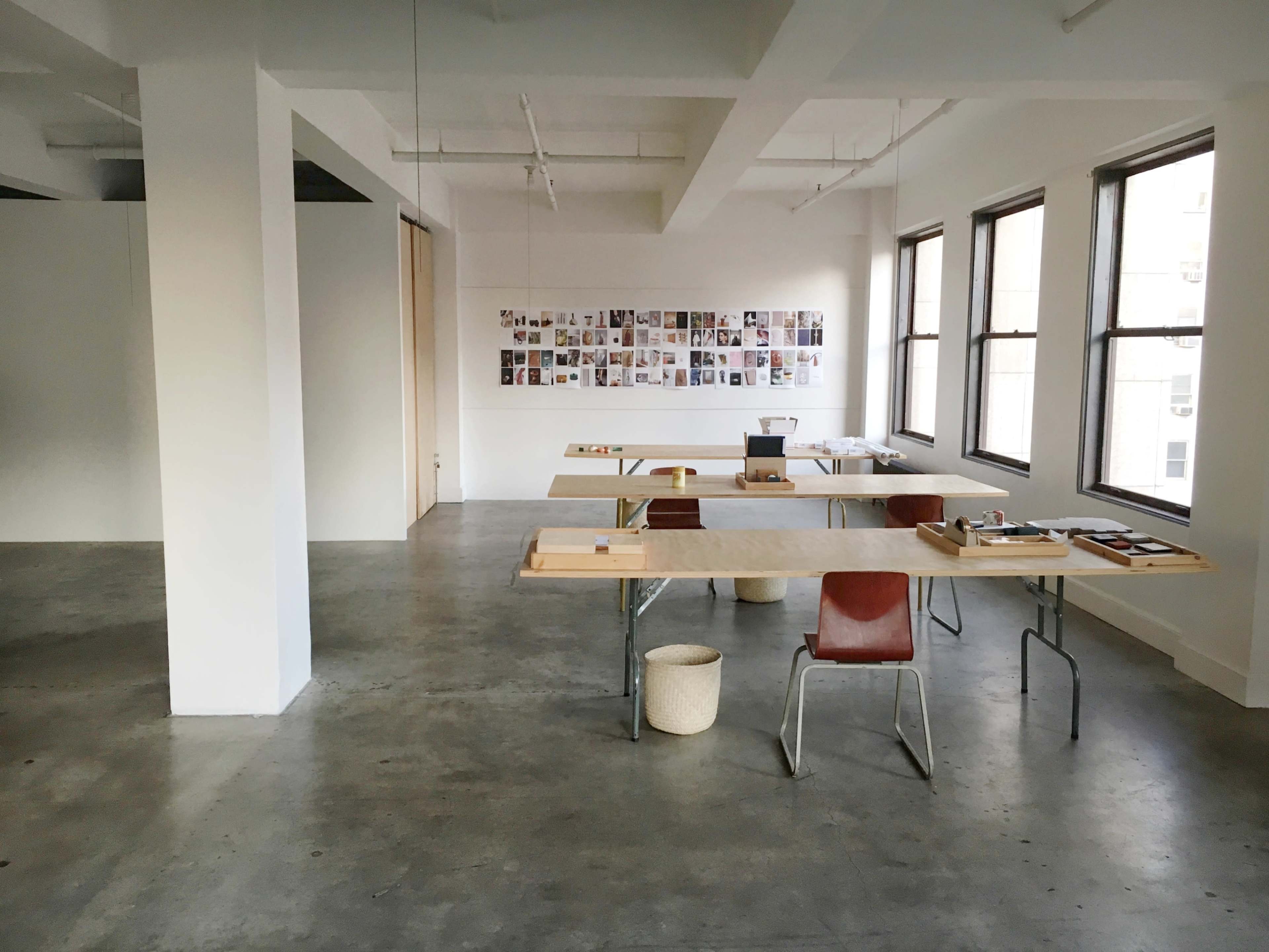 An empty, minimalist workspace features wooden tables and chairs, with a wall adorned with a collection of photographs.