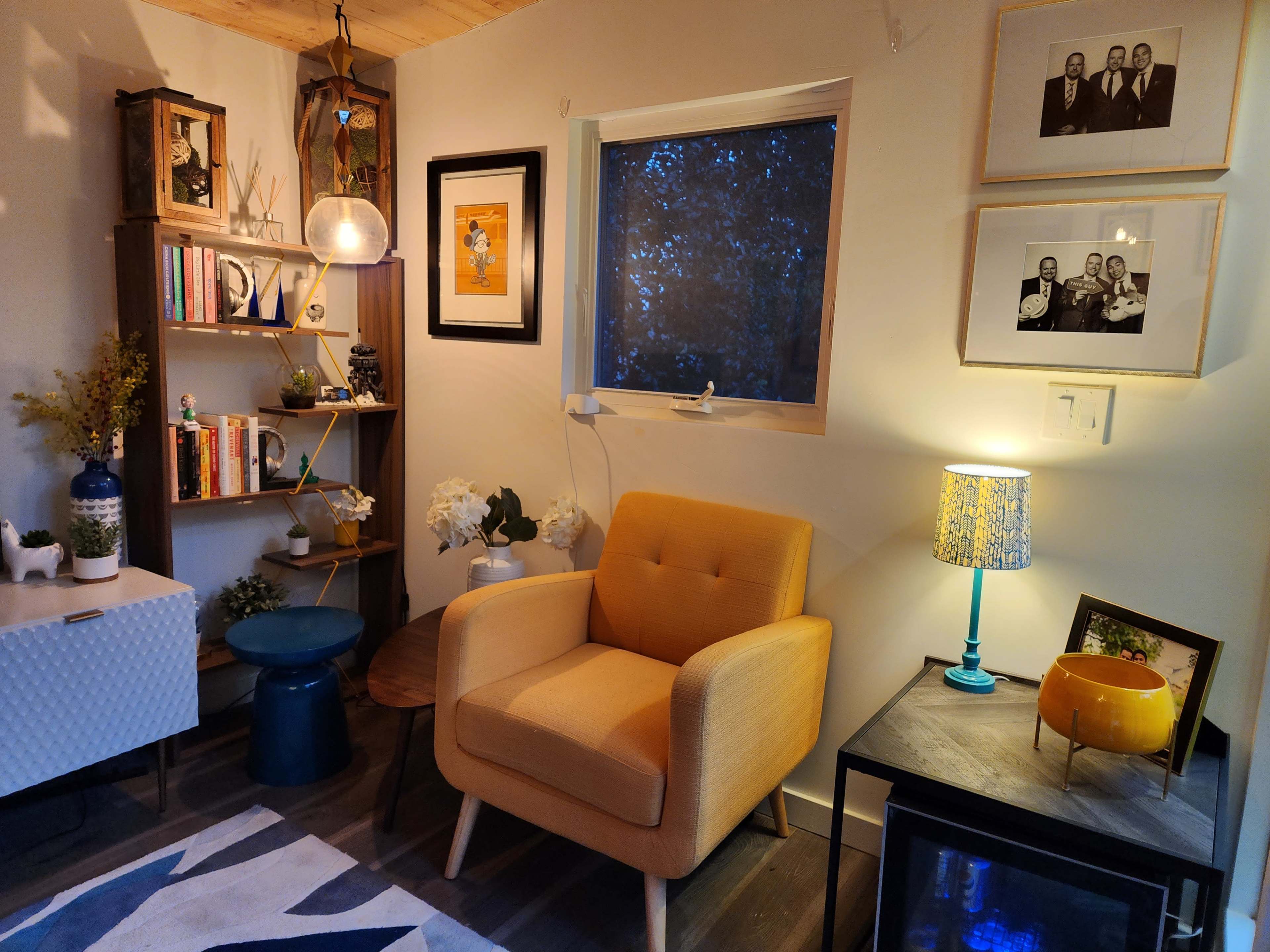 A cozy living room corner with a yellow armchair, bookshelf filled with books and decor, a side table, and a lamp.