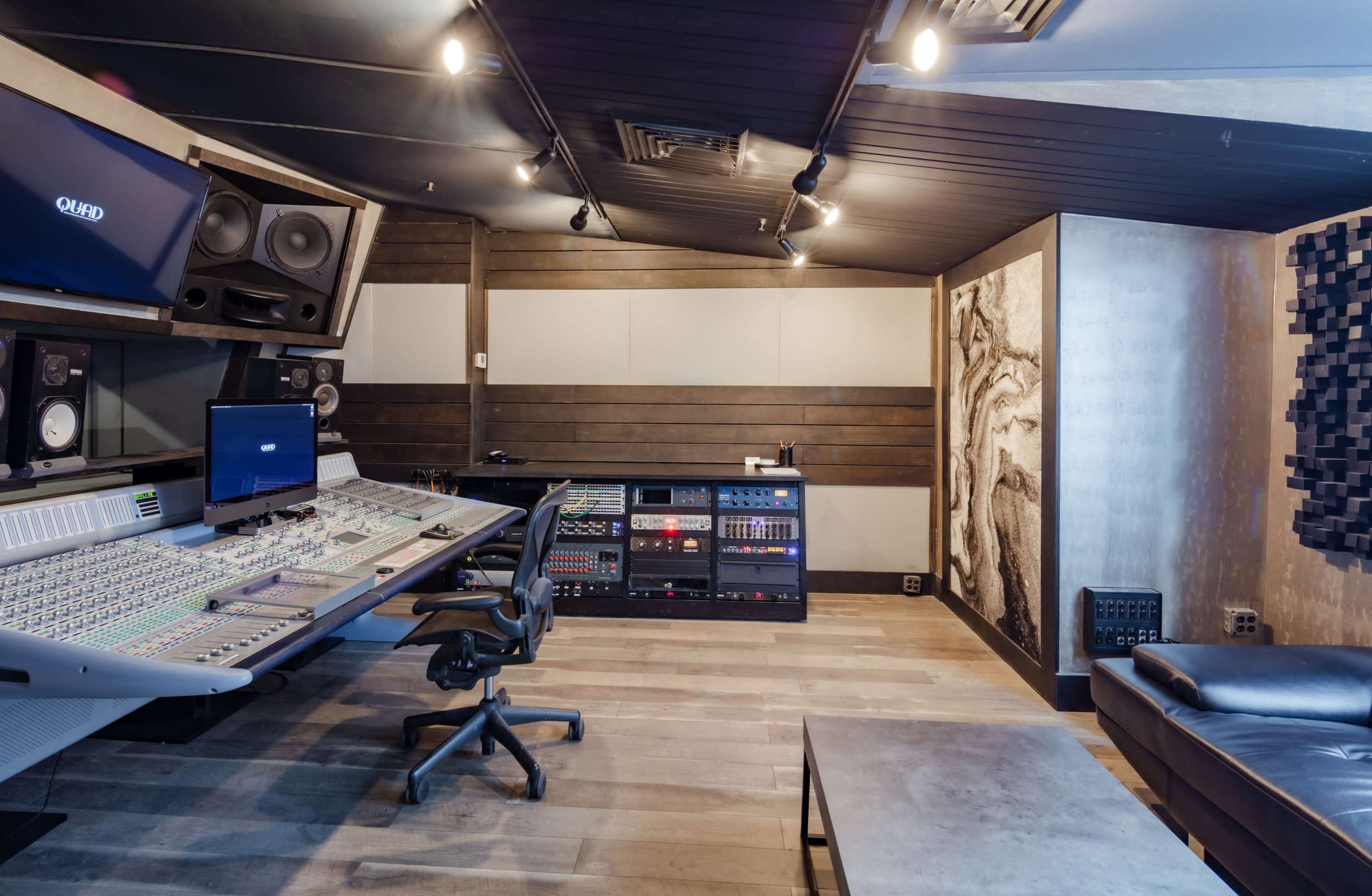 The image shows a modern recording studio featuring a large mixing console, computer monitors, and sound equipment against a backdrop of wooden panels and acoustic treatments.