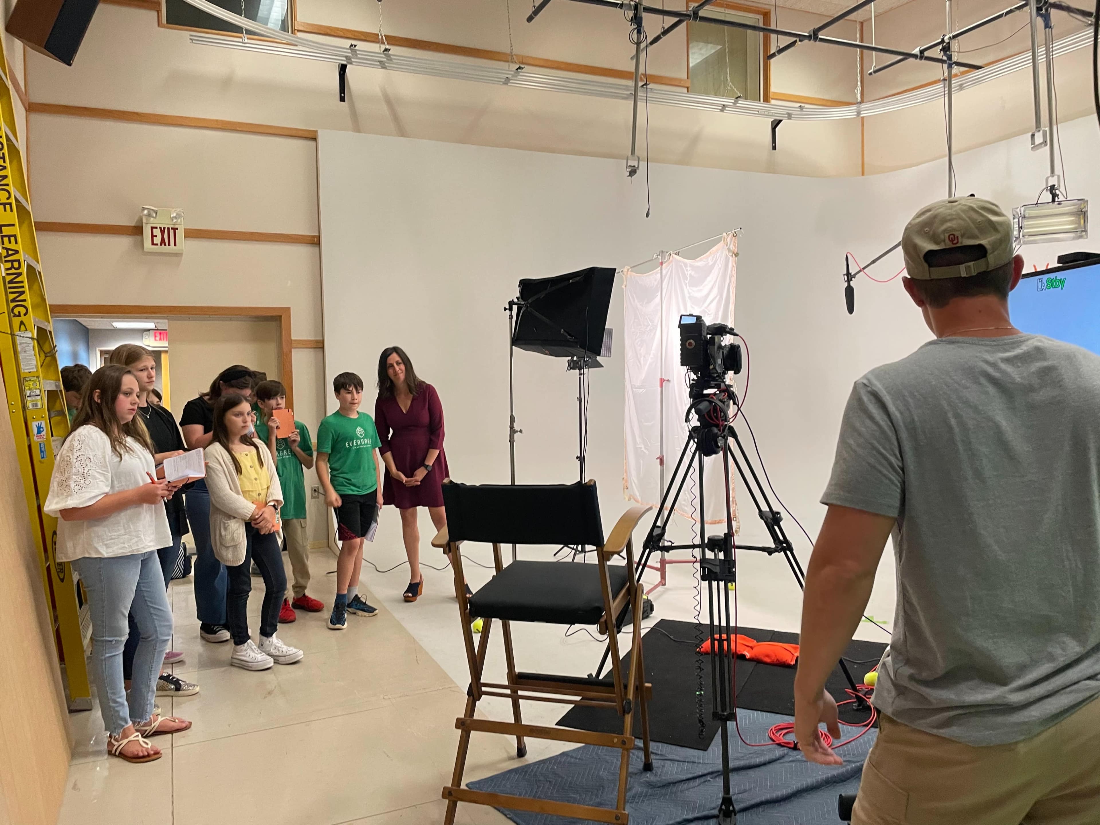 A group of six people stands in a studio while a man adjusts a camera on a tripod in front of them.
