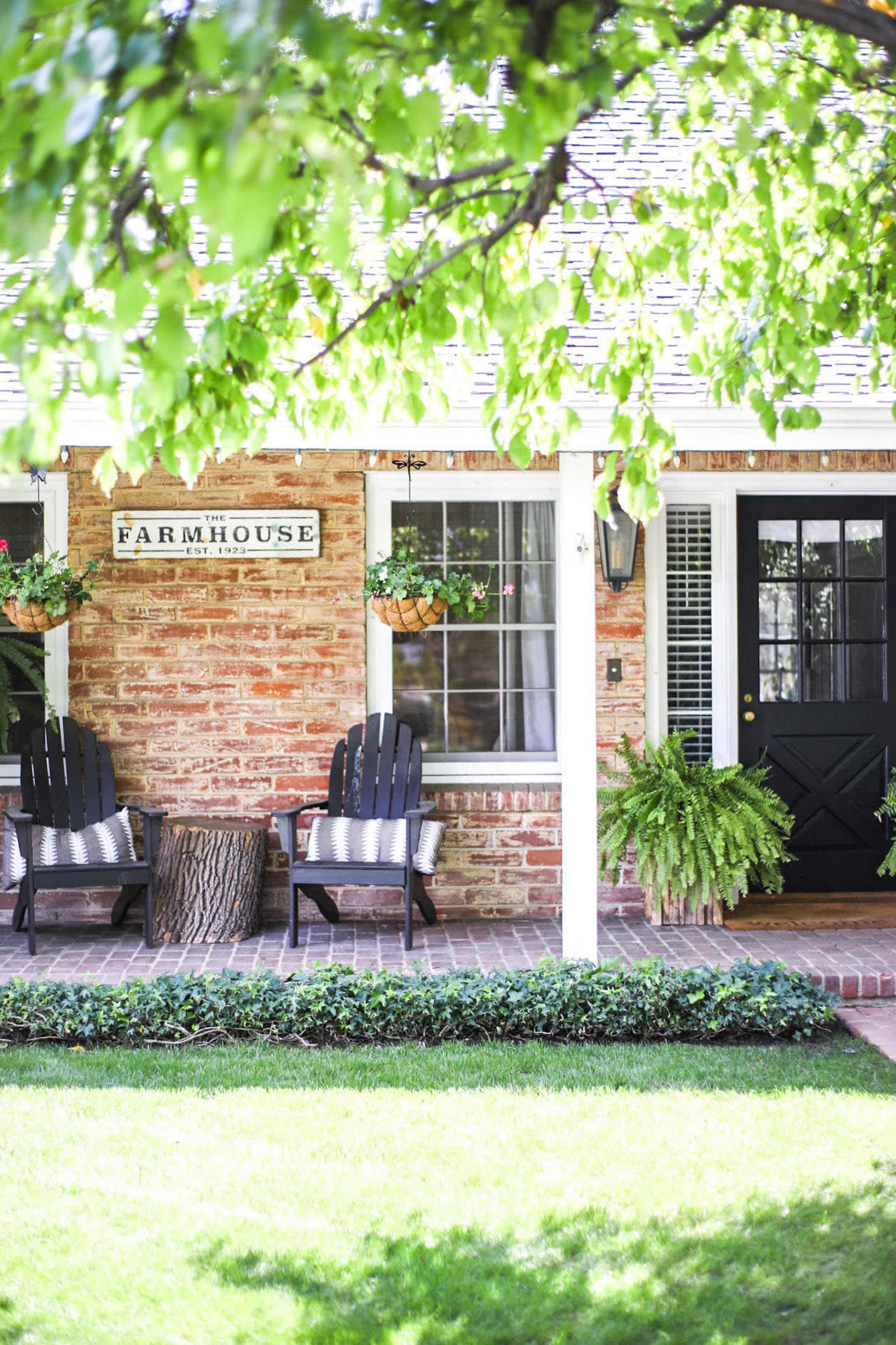 A brick farmhouse with a porch featuring two black chairs, potted plants, and a grassy lawn.