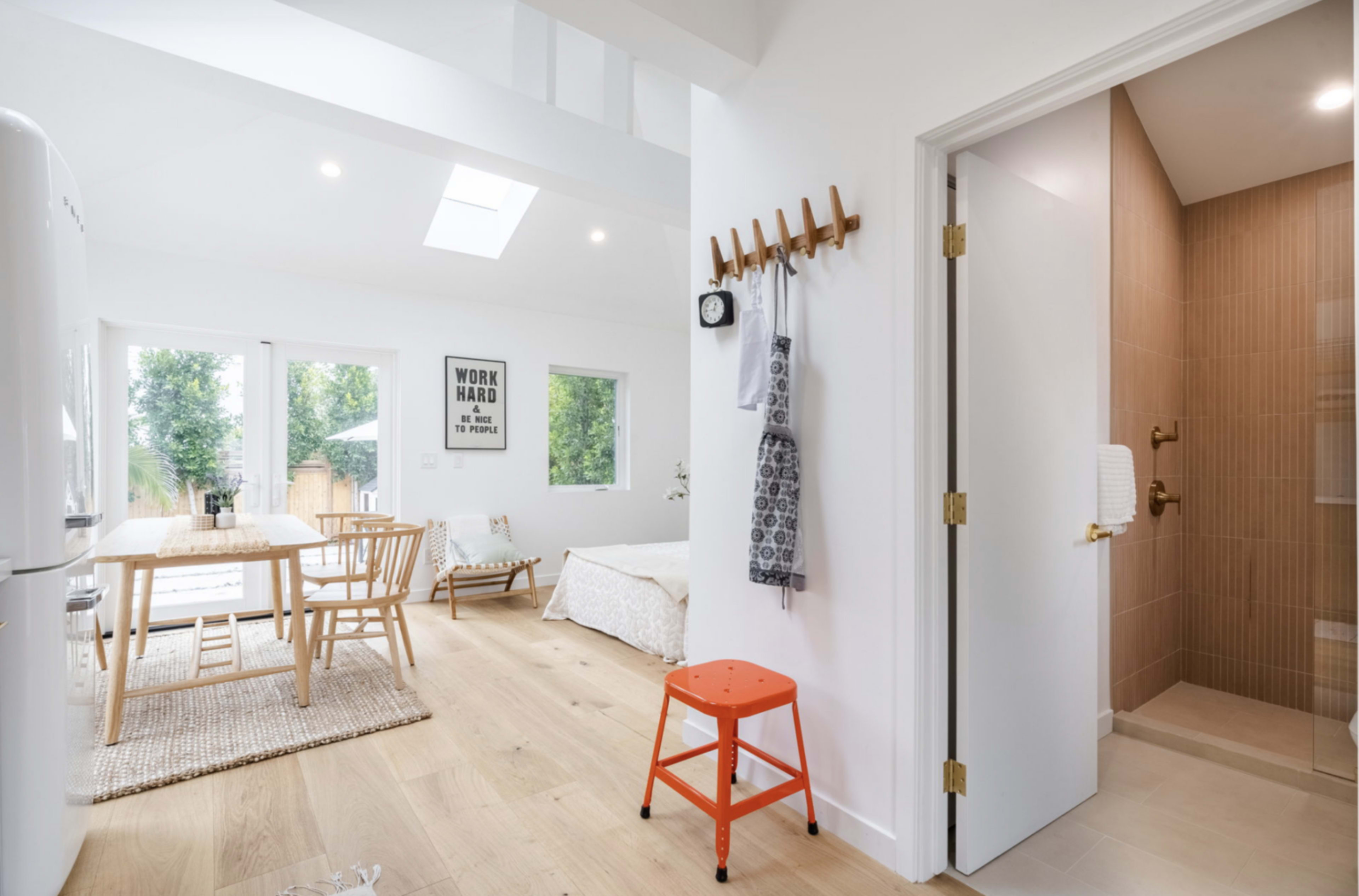 A bright, open interior space featuring a dining area with a wooden table, a kitchenette, and a bedroom area, with a bathroom visible through a door on the right.