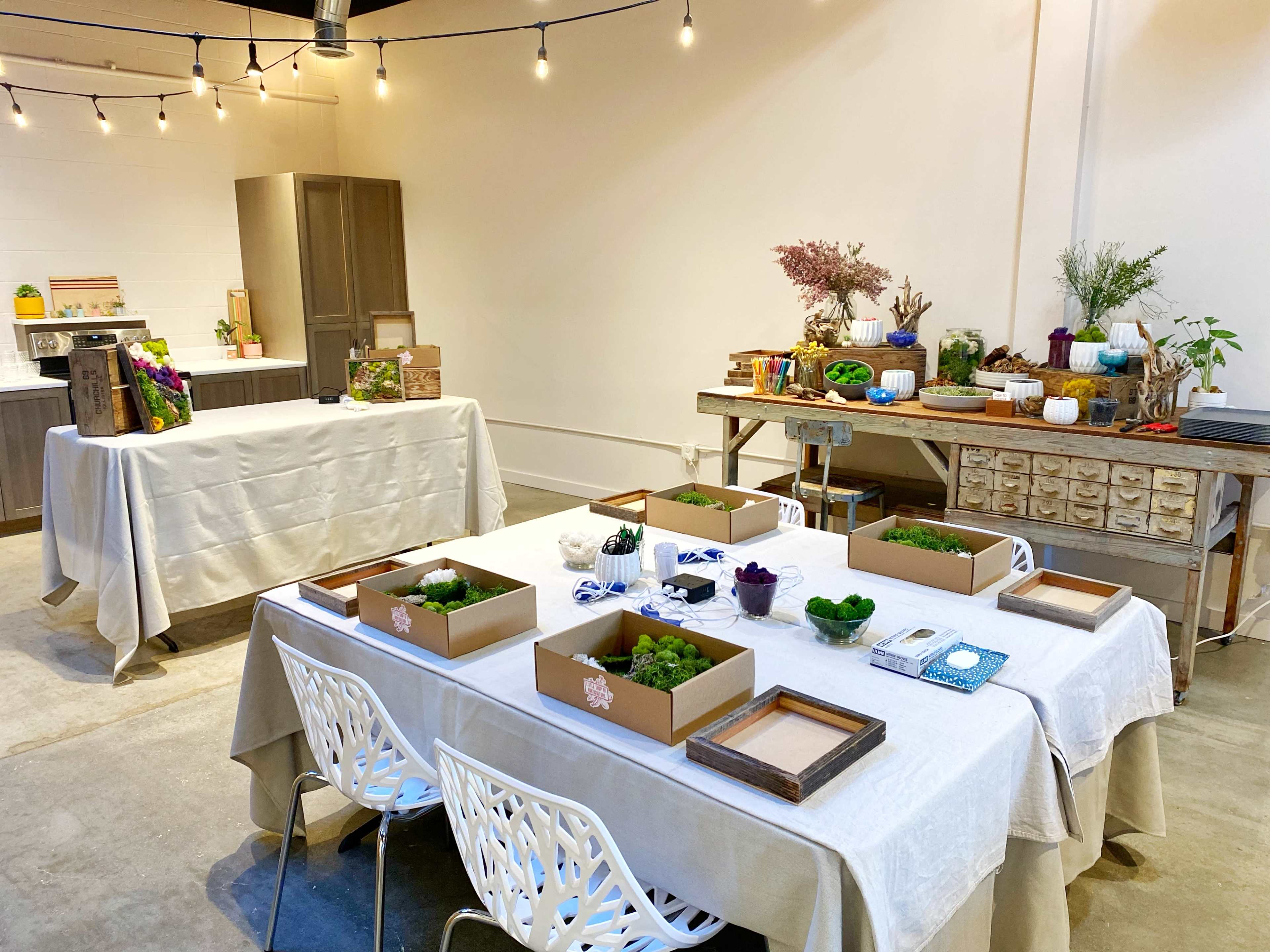 A bright, organized workshop space with multiple tables set up, each featuring containers of greenery and decorative items, along with a kitchen area in the background.