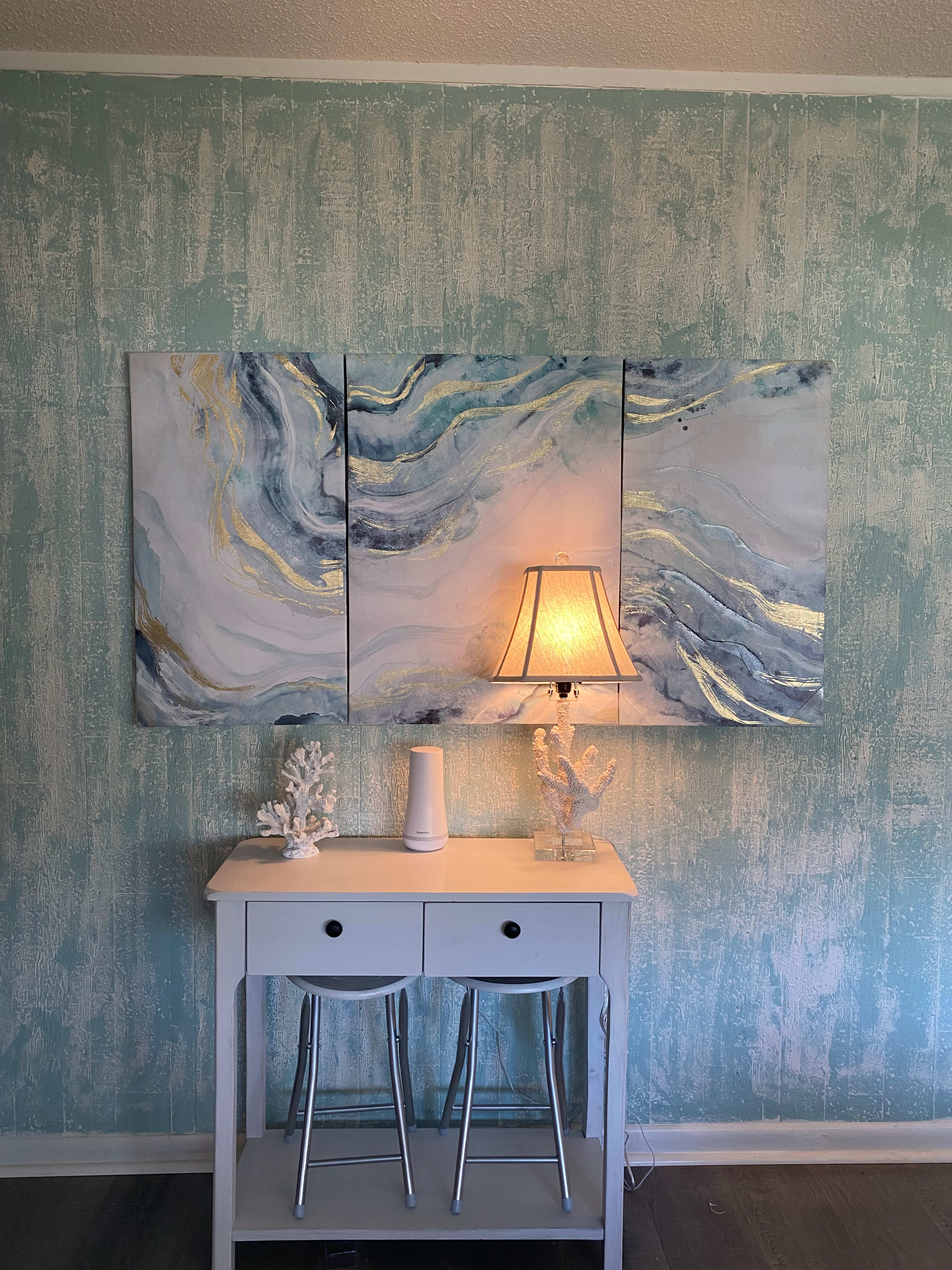 A light-colored table with two stools beneath a triptych painting of swirling patterns, accompanied by a lamp and decorative coral pieces against a textured blue wall.